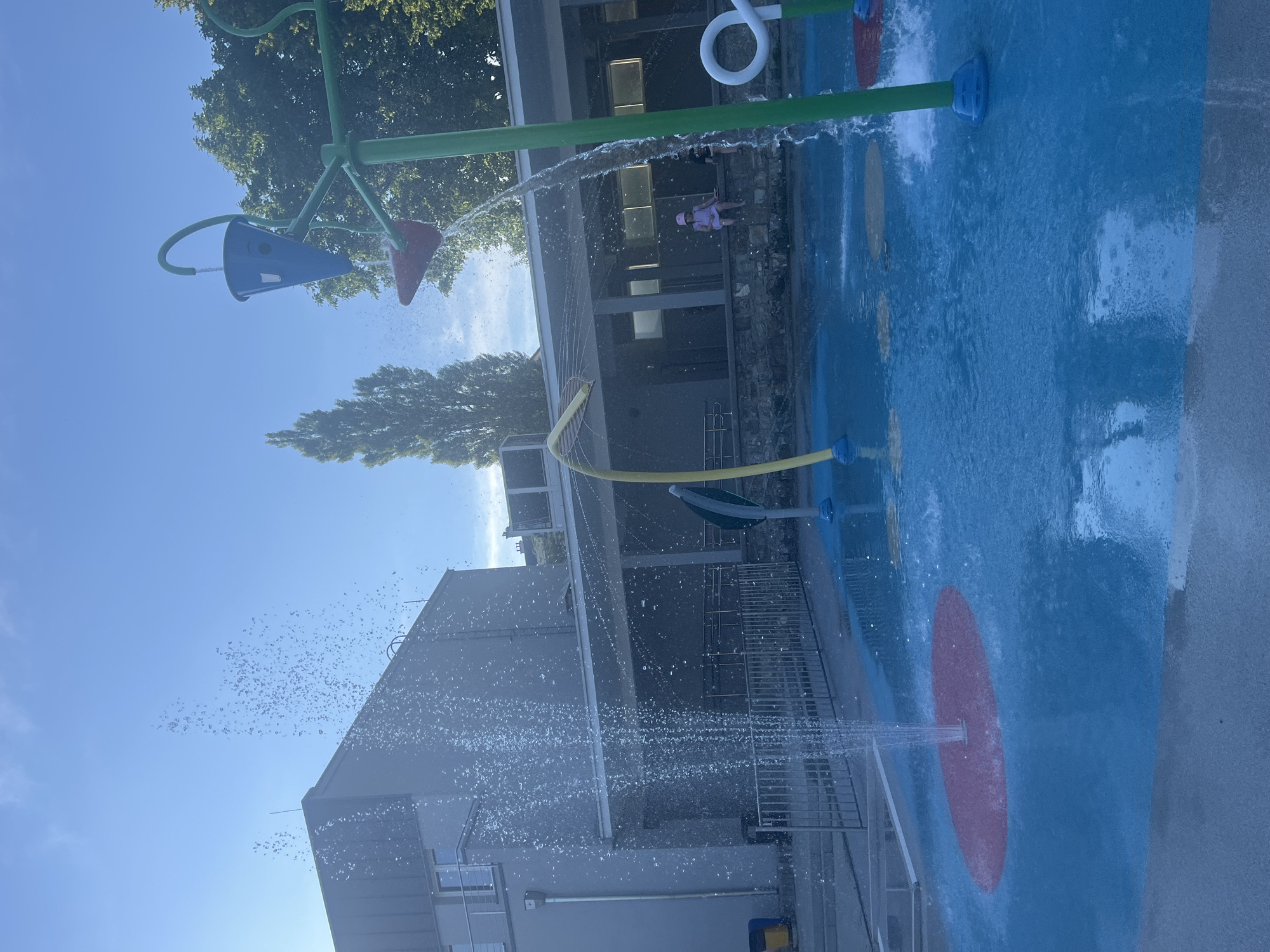 An outdoor splash pad is depicted on a sunny day under a clear blue sky. The splash pad features bright blue surfacing with colorful circular inlays of red and yellow. Several water features are active, including a tall spray jet in the foreground, a tipping bucket on a green pole, and various other spouts and sprayers. In the background, a modern building with large windows and a grey facade is visible, along with trees and a wall. Two children are seated on the wall.