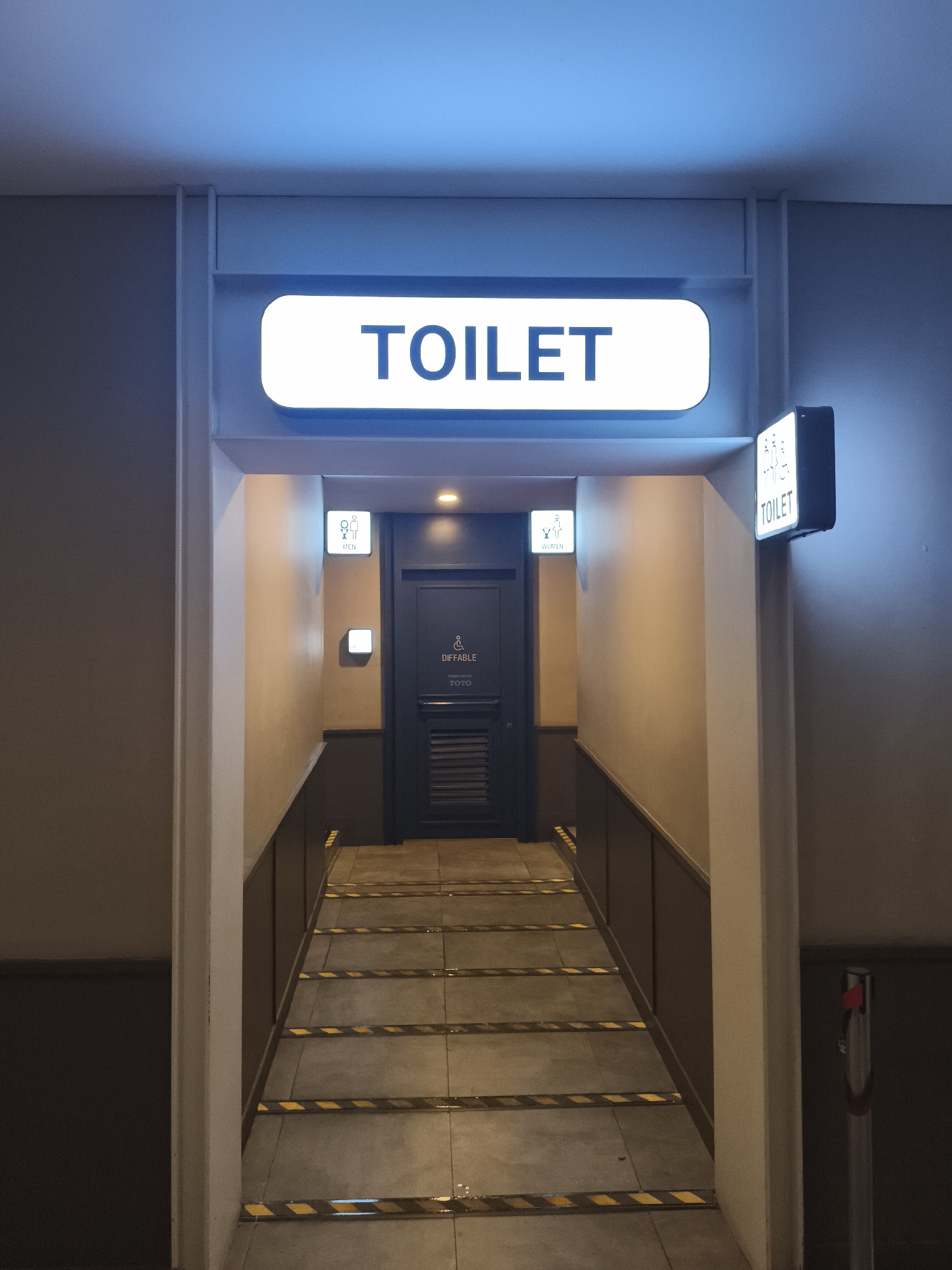 The image depicts an indoor hallway leading to a restroom. A large illuminated sign at the top reads "TOILET" in blue sans-serif font on a white background with rounded edges and a blue glow. To the right, a smaller illuminated sign displays "TOILET" above restroom icons for men and women. Further down the hallway, on the left, a sign with a female figure and "WOMEN" indicates the women's restroom. On the right, a sign with a male figure and "MEN" indicates the men's restroom. Beyond the "MEN" sign, a dark door is visible with a disabled symbol and "DIFFABLE" text above "TOTO," suggesting an accessible restroom. The hallway floor is tiled with yellow and black striped caution tape marking the edge of the steps. The walls are painted in neutral tones, with the hallway illuminated by overhead lights and the signs. The overall scene appears to be in Depok, Indonesia, indicated by the presence of accessible restrooms often found in public places there.