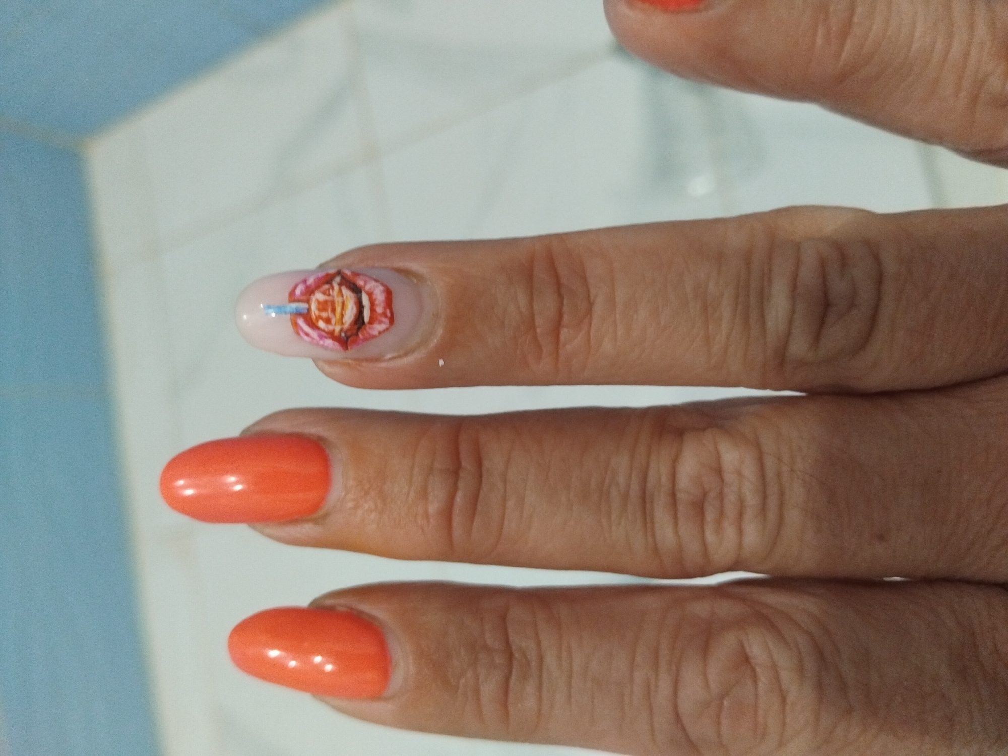 The image shows a hand with three fingers visible. Two of the fingernails are painted orange. The fingernail of the index finger has a light pink base and a design featuring a red and orange flower-like pattern with a blue horizontal line across the middle. The setting appears to be indoors, with a white and light blue tiled background. The image does not provide information about the location in the city of Stara Zagora, Bulgaria.