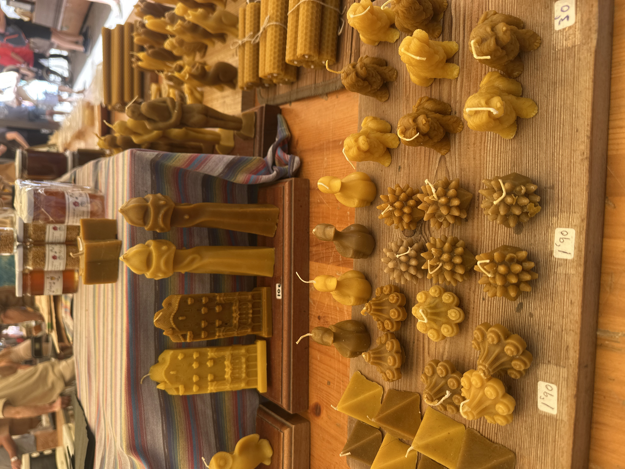 This image depicts a close-up, eye-level view of a market stall showcasing an assortment of beeswax candles, likely located in Barcelona, Spain, as evidenced by the general atmosphere of a European outdoor market. The stall is adorned with a striped cloth, and the foreground is dominated by various intricately shaped beeswax candles displayed on wooden planks.
In the foreground, an array of small, decorative candles are arranged. These include pyramid-shaped candles, several candles shaped like fluffy dogs, and numerous starburst or floral-shaped candles. Some of these candles have small white price tags attached, with visible prices like "1.90".
Further back, a collection of taller, more elaborate beeswax candles are visible. These are shaped like buildings, a person holding another person, and abstract pillar forms. Some of these are placed on wooden stands. Behind these, a stack of cylindrical beeswax candle rolls with a honeycomb pattern are neatly arranged.
The background is blurred but suggests a bustling market environment. There are glimpses of people in casual attire, some interacting with the stalls or walking through the market. The lighting suggests it is daytime, possibly outdoors or in a well-lit indoor market. The overall impression is one of a craft or artisanal market offering handmade goods.
