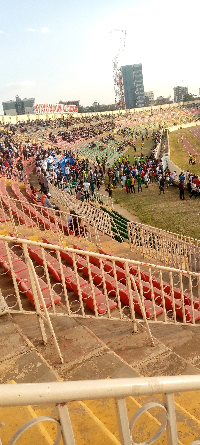 An outdoor stadium with tiered seating colored red, yellow, and green is filled with spectators. People gather on the green field next to the seating area. A tall metal tower stands in the background, adjacent to a tall building. A banner reads "NYAYO NATIONAL STADIUM" across the top of the stadium. The image is taken at an upward angle from the lower seating area, offering a partial view of the surroundings.