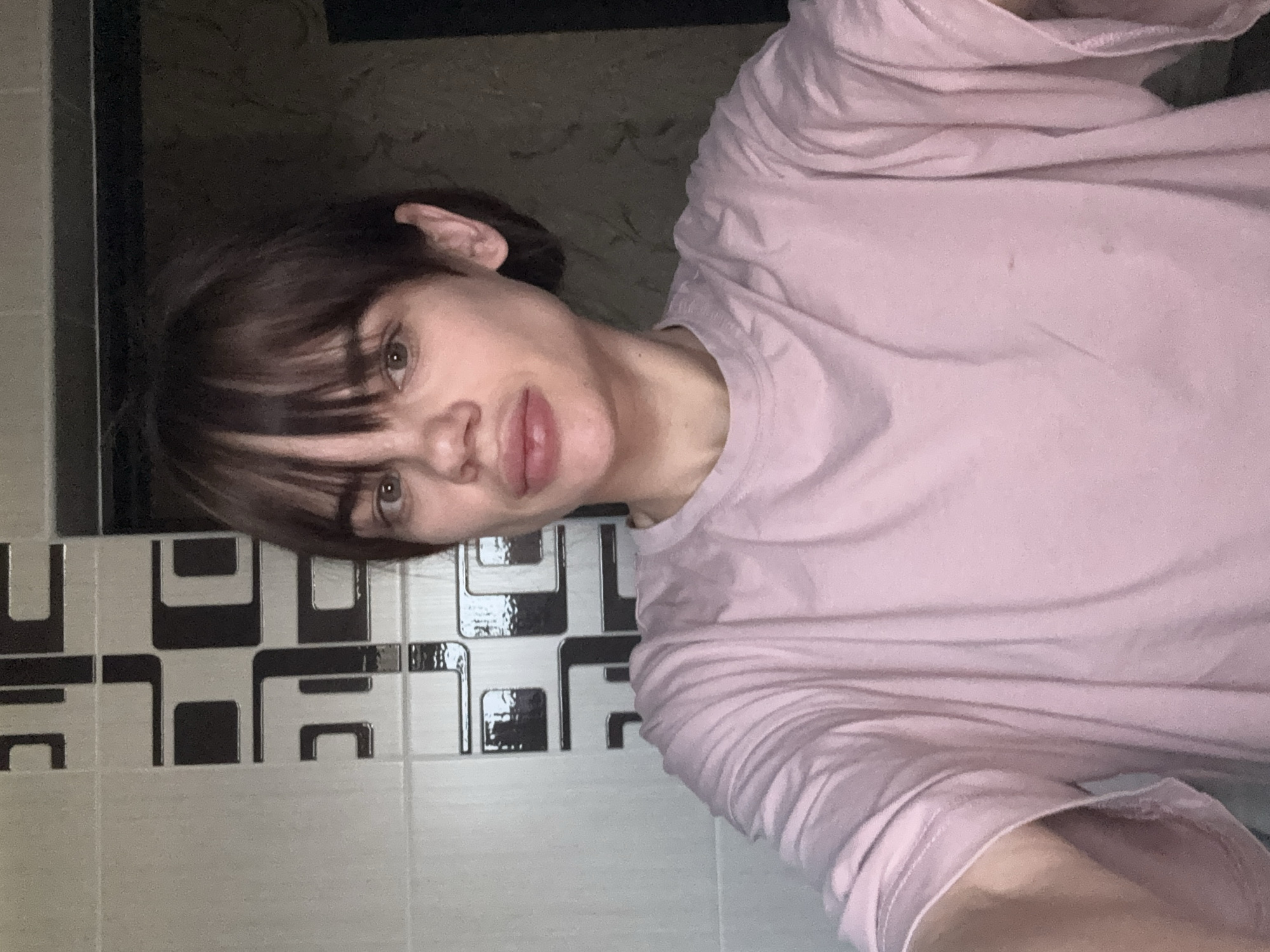 A person with short, dark hair and bangs, wearing a pink t-shirt, is positioned in front of a tiled wall. The tiles are light with a repeating black and white geometric pattern. The background features a dark, undefined area. The photograph appears to be taken in a bathroom setting. The location in the city of Rostov-on-Don, Russia, is not discernible from the visual information provided.