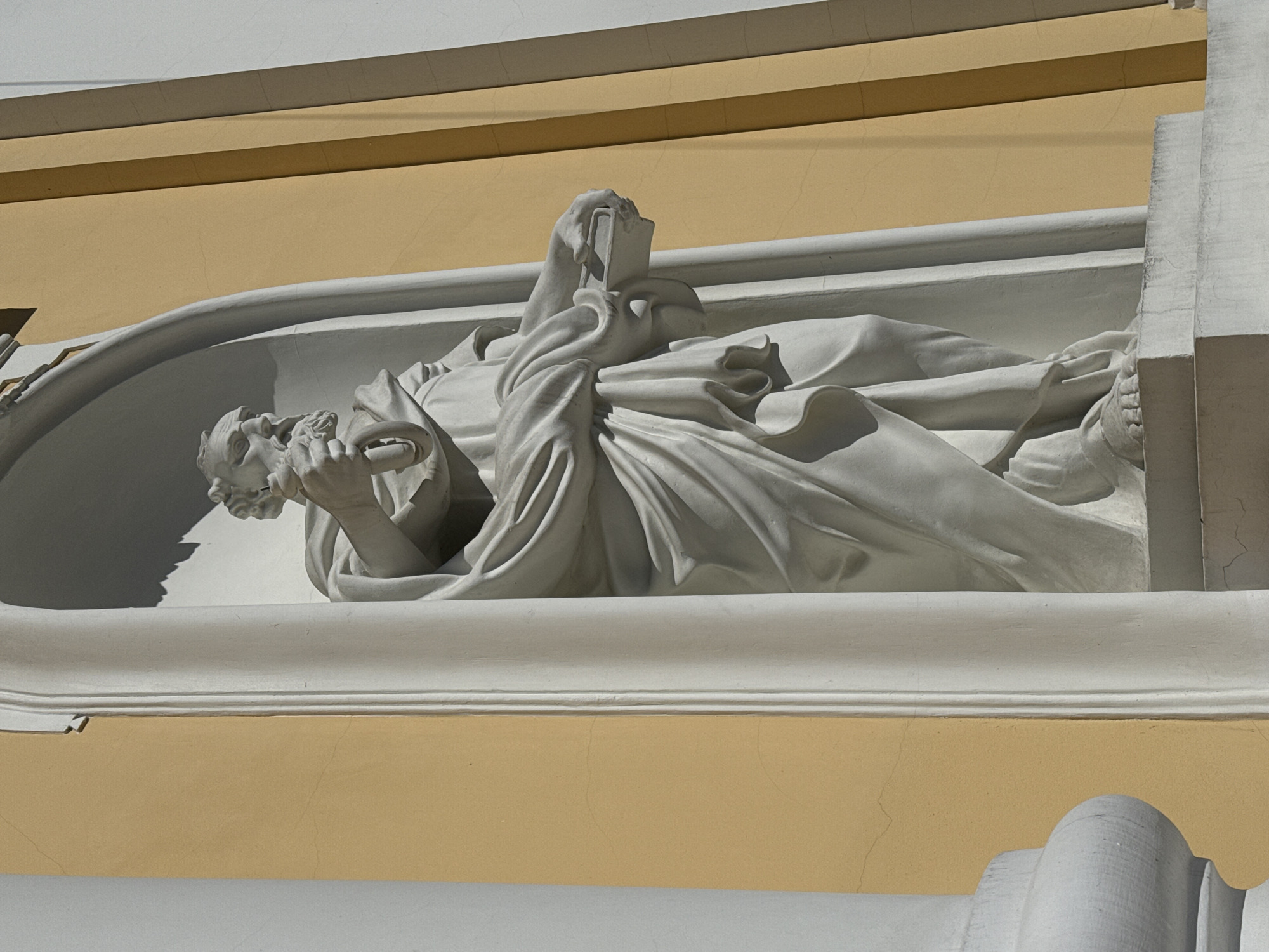 The image captures a white, high-relief sculpture integrated into the facade of a building, likely in Saint Petersburg, Russia. The sculpture depicts two figures within a shallow, curved niche.
The central figure is a man lying supine, partially covered by flowing drapery. His head is slightly raised, and his left hand, holding an object that resembles a scroll or a thick book, rests near his chest. His expression is not clearly discernible but suggests repose or deep contemplation.
Above and slightly draped over the man is a second figure, likely female. Her head is bowed and obscured by her arm and drapery as she holds an open book in her hands. Her posture conveys a sense of sorrow, lamentation, or deep study, possibly in mourning or remembrance of the man below. The flowing garments of both figures are intricately carved, creating strong shadows and highlights that emphasize their forms.
The sculpture is set against a pale yellow (ochre) wall of the building, which shows minor cracks, indicating age. Above and below the niche are architectural elements painted white, providing a stark contrast to the yellow wall. The bright sunlight, likely from the left, casts strong, well-defined shadows, especially on the recessed parts of the sculpture and the wall, indicating a clear, sunny day. No visible text is present on the sculpture or the building. The overall style suggests classical or neoclassical influences, characteristic of historical architecture in Saint Petersburg.