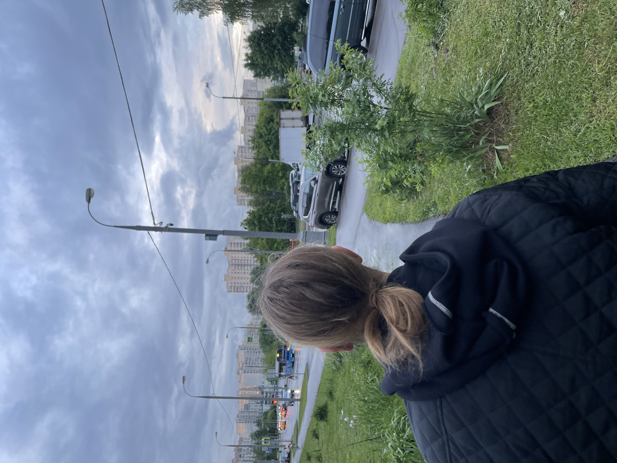 The image shows a person with light brown hair in a ponytail, wearing a black quilted jacket with a hood. The person is standing near a sidewalk with greenery on the right. Beyond the sidewalk is a road with streetlights, traffic, and buildings in the distance, indicating a city environment. The sky is overcast. Based on the architecture and general appearance, the location appears to be Moscow, Russia. Several cars are visible along the road and parked on the side of the street.