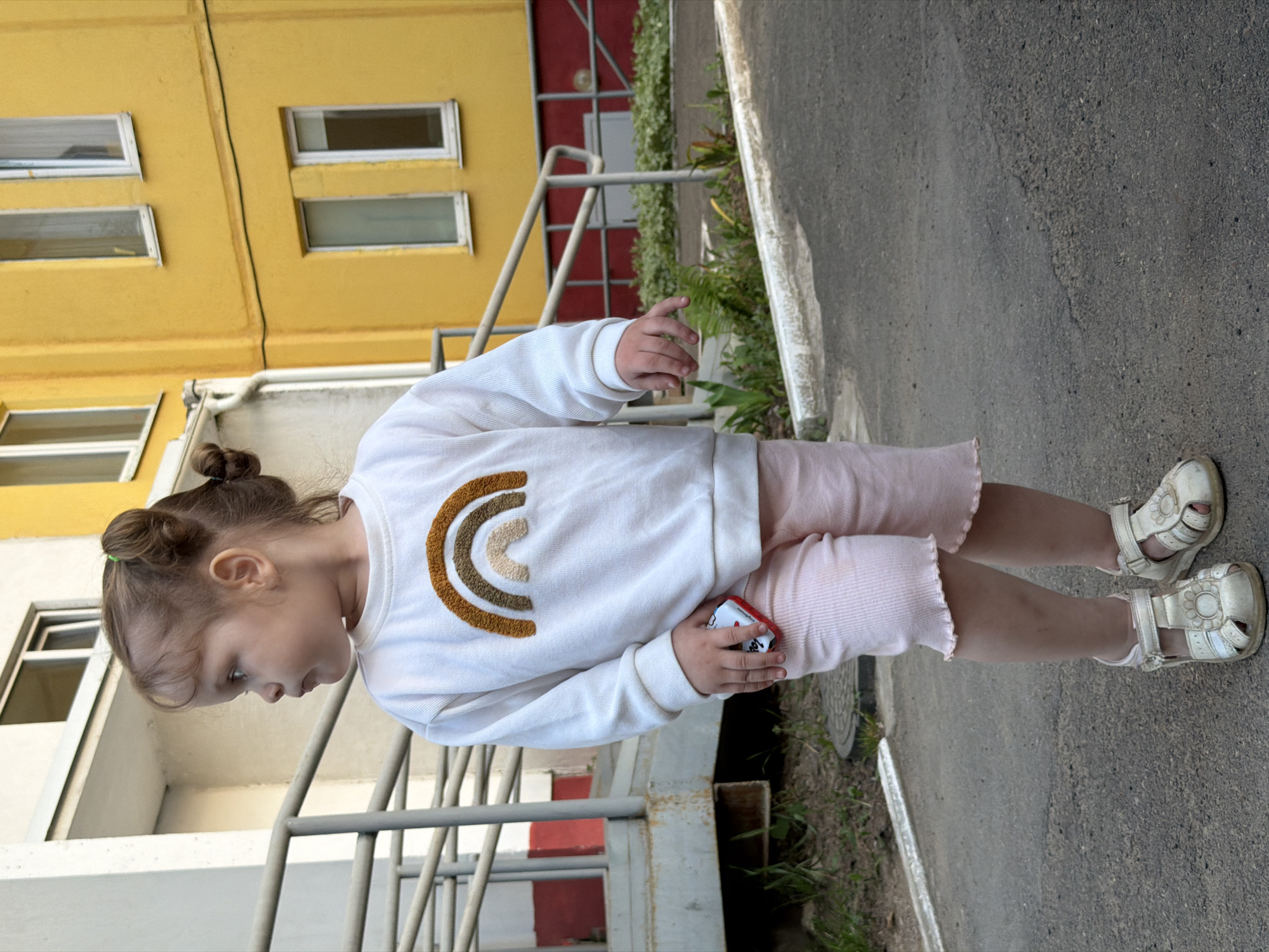 A toddler girl stands on a grey asphalt surface outdoors. She is wearing a white long-sleeved shirt with a brown and beige rainbow design, light pink ruffled shorts, and white sandals. She has brown hair pulled into two small buns. She is holding a small red and white object in her right hand.
Behind the girl is a metal staircase railing with red accents. Behind the staircase is a multi-story yellow building with white-framed windows. Patches of greenery are visible along the edge of the asphalt. The scene is located in the city of Chaikovsky, Russia.
