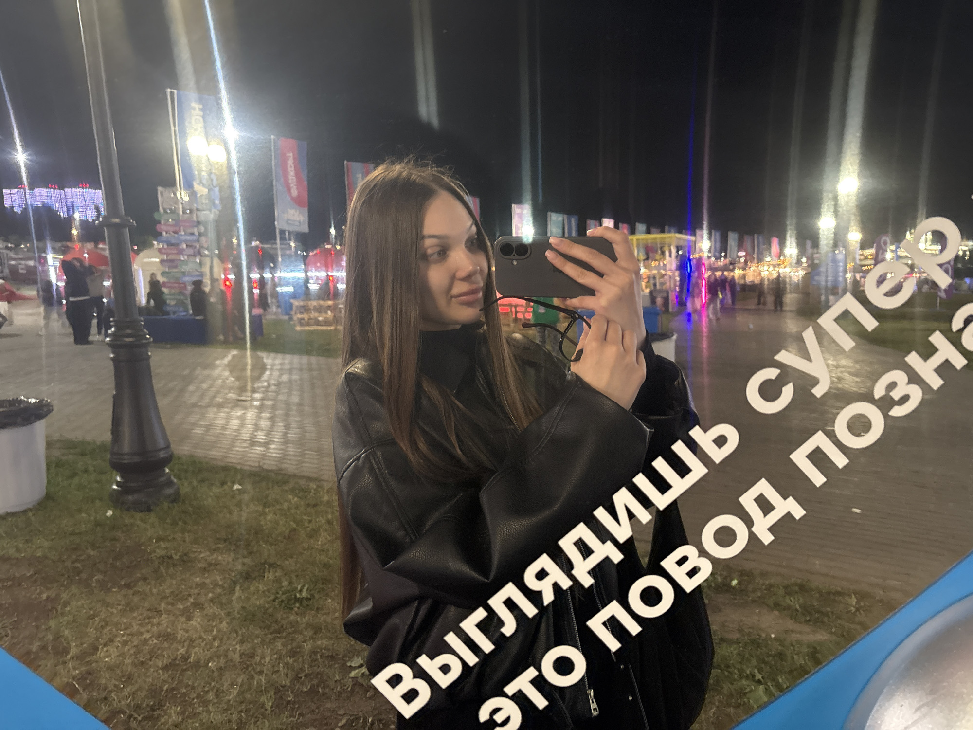 This image depicts a young woman taking a selfie at night in a reflective surface, likely glass or a mirror, located in an outdoor urban public space in Kazan, Russia.
The central figure is a young woman with long, straight, dark brown hair. She is wearing a black leather jacket over a dark, possibly high-necked top. She holds a dark-colored smartphone, likely an iPhone with two visible camera lenses, in her right hand, pointed towards her reflection. Her left hand holds a pair of dark-rimmed glasses. She has a slight smile or smirk on her face and is looking towards the phone's screen.
The image is a reflection, indicated by the mirrored text visible on the surface. The white Cyrillic text, when read correctly (un-mirrored), states: "ВЫГЛЯДИШЬ СУПЕР ЭТО ПОВОД ПОЗНАКОМИТЬСЯ", which translates to "YOU LOOK SUPER THIS IS A REASON TO MEET" or "YOU LOOK SUPER THIS IS AN OCCASION TO GET ACQUAINTED".
The background, also a reflection, shows a bustling outdoor environment illuminated by numerous artificial lights. Multiple tall streetlights with bright, star-like glows and light streaks are visible. Blurred figures of people can be seen walking in the distance, suggesting a public gathering or event. Various illuminated structures, possibly market stalls or pavilions, with colorful lights (red, blue, yellow) are present. Several vertical banners are also visible; one prominent blue banner on the left displays "HOKA" in white text, and a red banner further right shows text that appears to be "ТРОНАВО" (TRONAVO) in white. The ground consists of both paved areas and patches of grass. A dark lamppost base and a white trash can with a black rim are visible in the lower-left corner. The bottom edge of the frame shows a blue border, likely part of the structure containing the reflective surface.