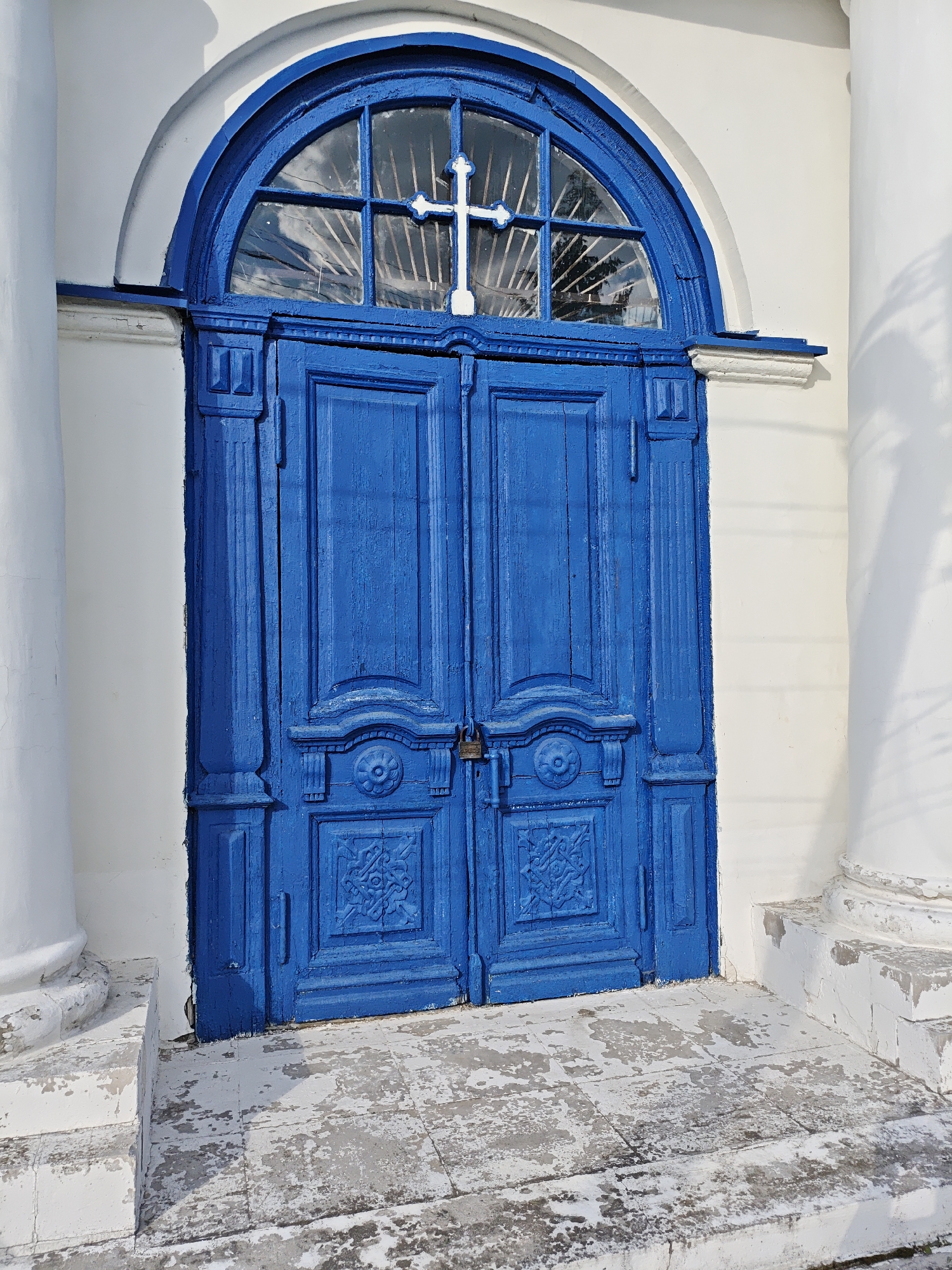 A blue arched double door with a cross in the transom stands at the entrance to a building in Мамонтово, Russia. The door has decorative carvings and is secured with a padlock. The building is white, with fluted columns on either side of the entrance. The ground is a worn, tiled surface. The scene is sunlit.