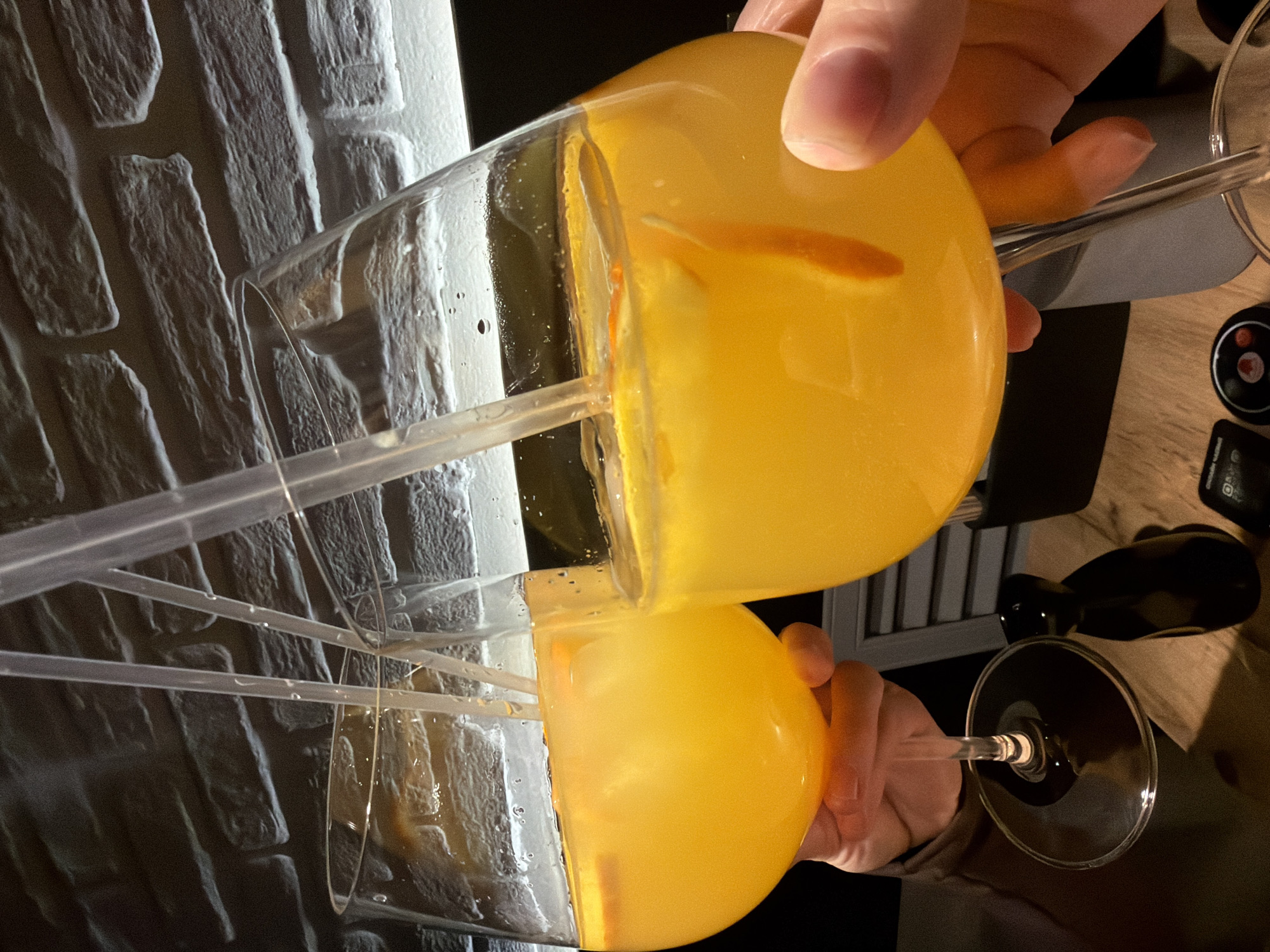 A person is pouring an orange-colored liquid from a large glass into another large glass, both containing straws and orange slices. The scene appears to be indoors. A brick-like patterned wall is visible in the background. Other items, including what looks like a bottle and electronics, are on the table.