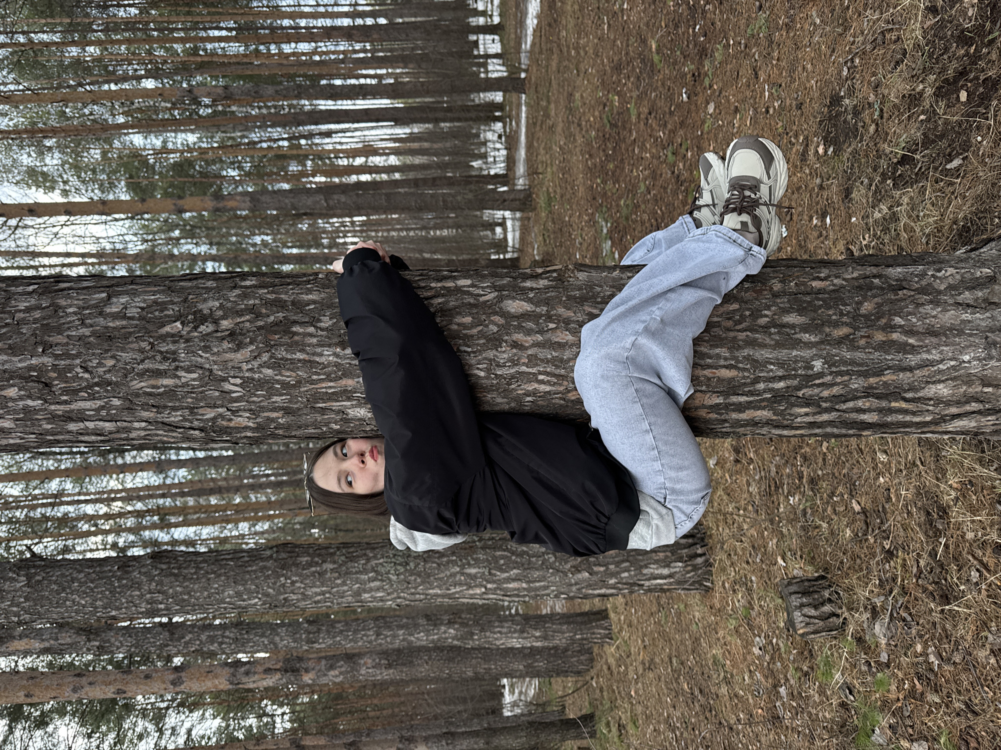 A young person, likely female, lies prone on a large fallen tree trunk within a pine forest. She wears light blue denim jeans, chunky athletic shoes in gray and beige, and a black windbreaker jacket over a gray hooded sweatshirt. Her arms grip the tree trunk behind her head. The forest floor is covered in brown pine needles and sparse undergrowth. Tall, straight pine trees dominate the background, appearing in rows. The lighting suggests an overcast day. The image orientation is rotated 90 degrees clockwise. The location is presumed to be in or around the city of Chaikovsky, Russia.