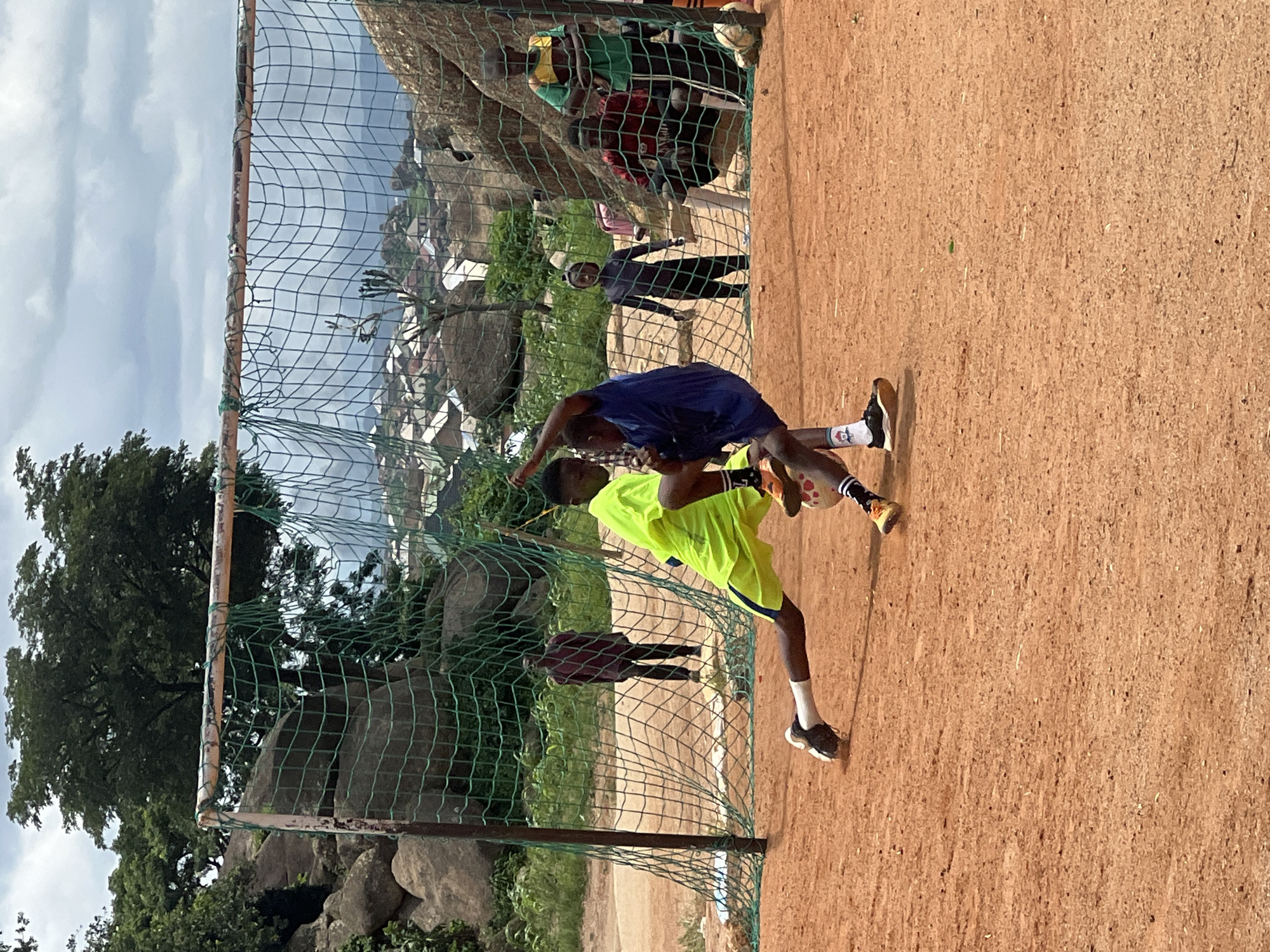 The image depicts an outdoor scene on a dirt field, likely in Jos, Nigeria, with a makeshift soccer goal. Two young males are actively engaged in a soccer game in the foreground. One player, wearing a bright neon yellow jersey and blue shorts, is on the ground, lunging forward with his right leg extended. The other player, in a dark blue jersey, is positioned behind him, also in motion, with his left arm raised. A soccer ball is visible between them. In the background, several other individuals are present, some observing the game and others standing near the goal. The environment features rocky outcrops and scattered vegetation, characteristic of the Jos Plateau. Beyond the immediate playing area, a settlement with houses and hills can be seen under a cloudy sky. The overall impression is one of a casual soccer match taking place in a rugged, natural setting.