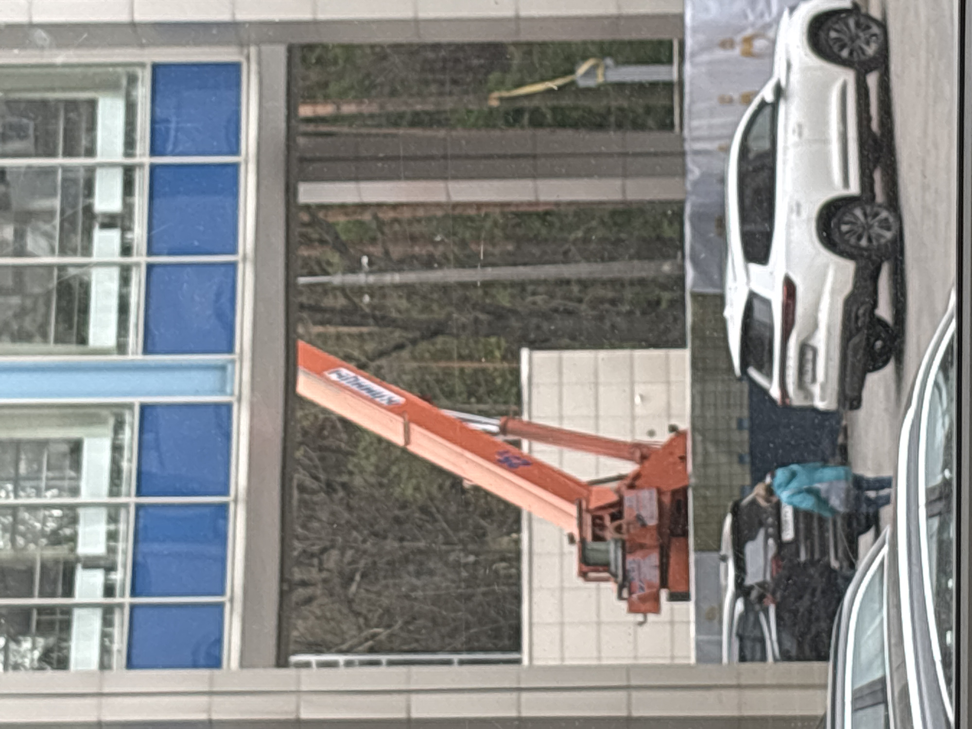 The image shows a reflection in a window. In the reflection, there is a white car parked on pavement, an orange aerial work platform, a building with blue windows, and several people. The location is Saint Petersburg, Russia.
