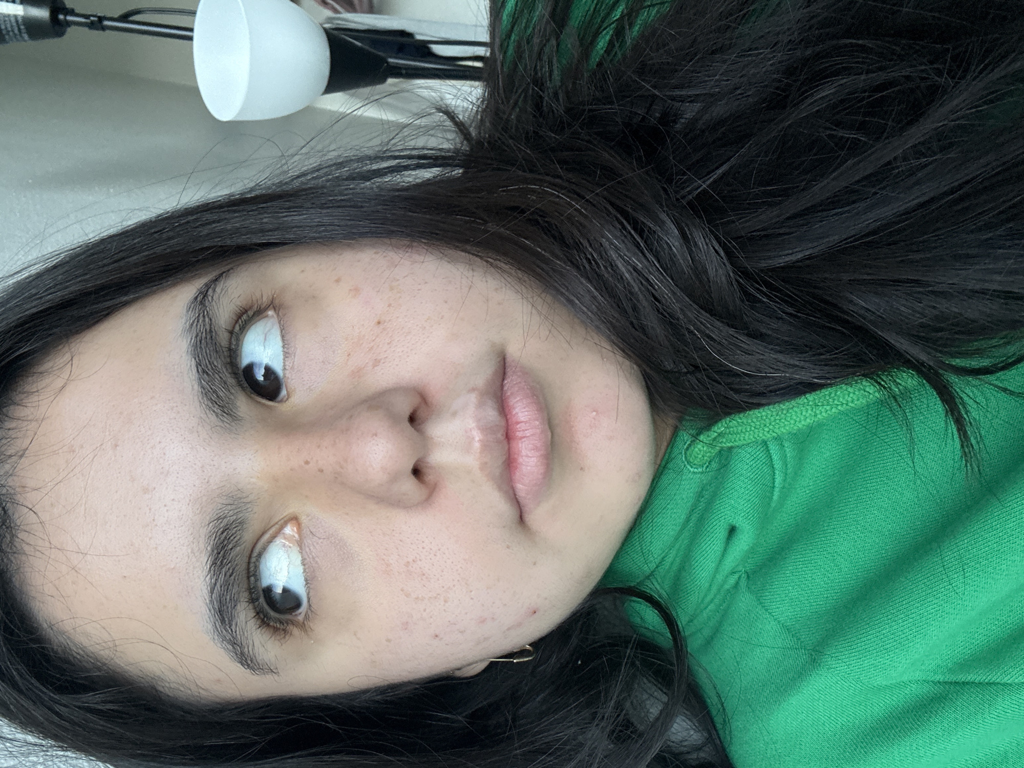 A close-up portrait shows a woman with dark hair and eyes looking upwards. She has fair skin with freckles and is wearing a green hooded sweatshirt. The background is blurred, but a white lamp fixture is visible in the upper left corner. No information about the location in Austin, Texas is discernible from the image.