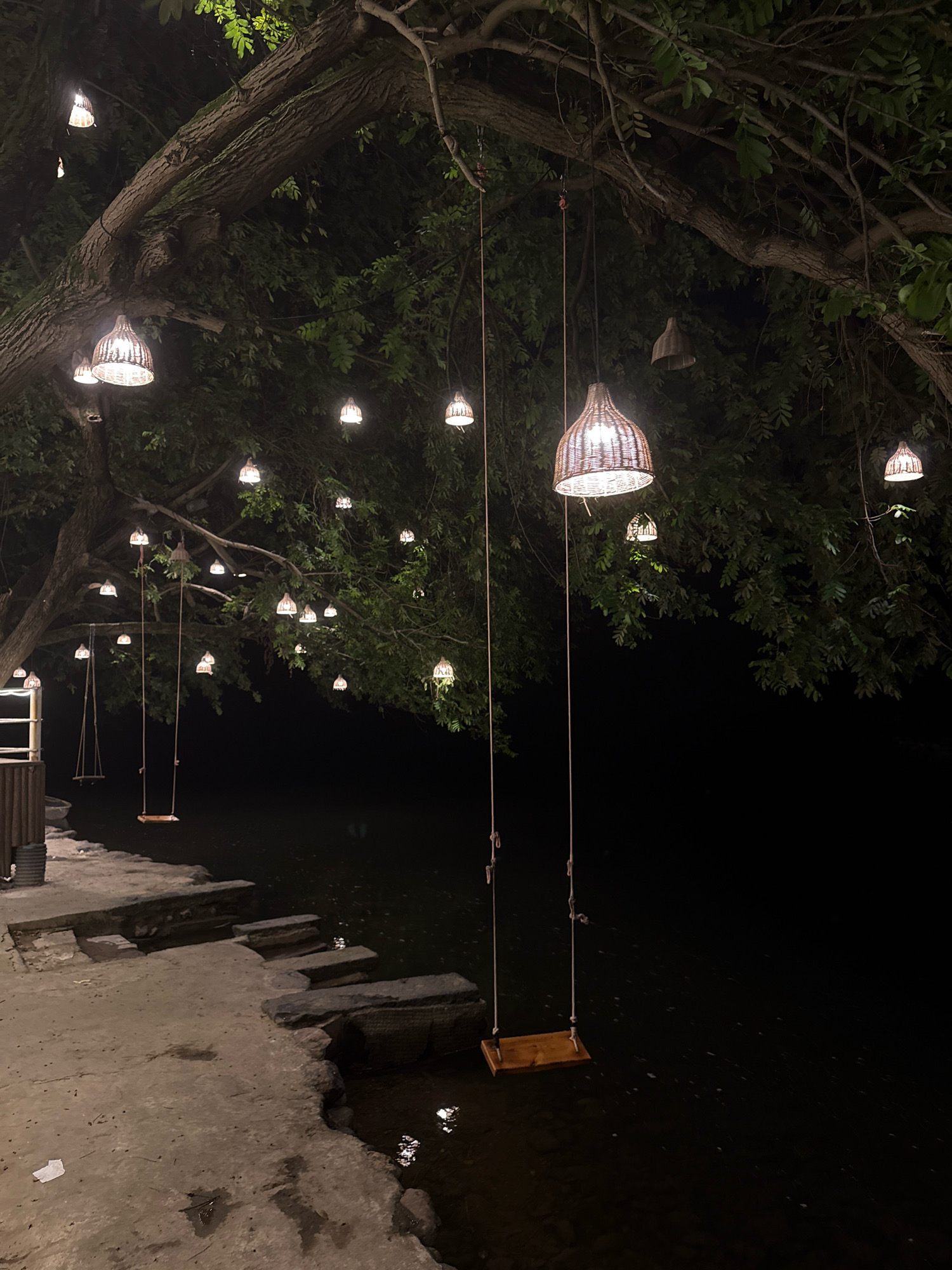 The image depicts an outdoor scene at night, illuminated by hanging lamps. These lamps, made of a woven material and shaped like inverted baskets, are suspended from the branches of a large tree. Several of these lamps are lit, casting a warm glow. Two swings are visible: one is a simple wooden plank suspended by ropes, hanging over dark water, and another appears to be located further back on a stone embankment. The area around the swings consists of a concrete or stone pathway with some rough-hewn stone steps leading down to the water. The water itself is dark and reflects some of the light from the lamps. The background is filled with the dark foliage of the tree and the blackness of the night sky. This scene is located in Yiwu, China, likely at a waterfront dining or recreational area. There are no people visible in the image.