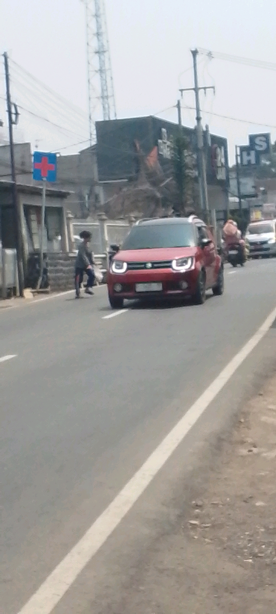 A low-angle, blurry, medium shot captures a street scene in Sukasari, Indonesia. In the foreground, the asphalt road is marked with a white line. A red car is positioned in the center, driving towards the viewer. To the left of the car, a pedestrian, appearing to be a child, is walking across the road. A red and blue sign with a white cross is visible on a pole near the pedestrian. In the background, various buildings, utility poles, and a tall tower can be seen. A motorcycle with a rider is visible in the background, to the right of the red car. The sky is overcast.