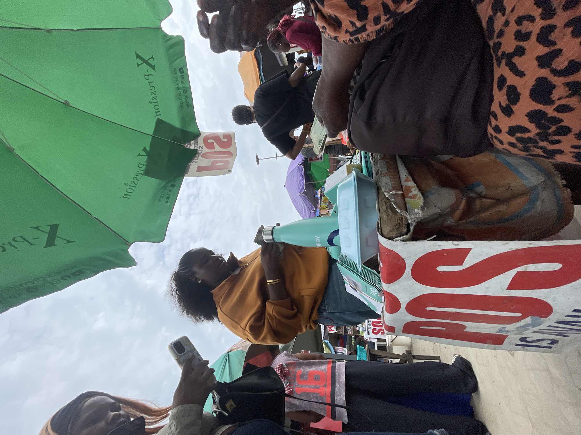 This is a low-angle, eye-level, outdoor photograph taken at a street-side market in Ajah, Nigeria. A woman in a yellow hooded sweatshirt and blue jeans is standing behind a table and appears to be handling cash. To her left, another person, mostly obscured, is looking at a smartphone. The table is covered with various items, including a light blue water bottle with "GoPay" branding, a light blue plastic container, and stacks of green pamphlets. A red and white sign with the letters "POS" in large red font and "IS AVAIL" in blue font is prominently displayed on the front of the table. Above the table, a large green umbrella with "X-Pression" branding is partially visible, providing shade. In the background, other market stalls, umbrellas, and people are present under a cloudy sky. A person in a black t-shirt is also visible in the middle ground, holding some cash. The overall scene depicts a commercial transaction or service being offered at a busy outdoor market.