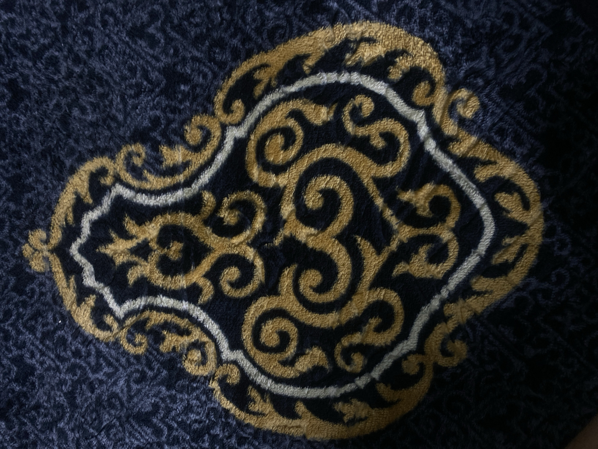 A close-up, overhead shot shows a dark blue, plush fabric with an ornate, symmetrical gold and white pattern. The pattern is centrally located and features swirling, arabesque-like designs. The background fabric has a subtle, repeating geometric texture. The image does not depict any identifiable urban environment or specific location within Alamüdün, Kyrgyzstan. There are no people, actions, or interactions visible.