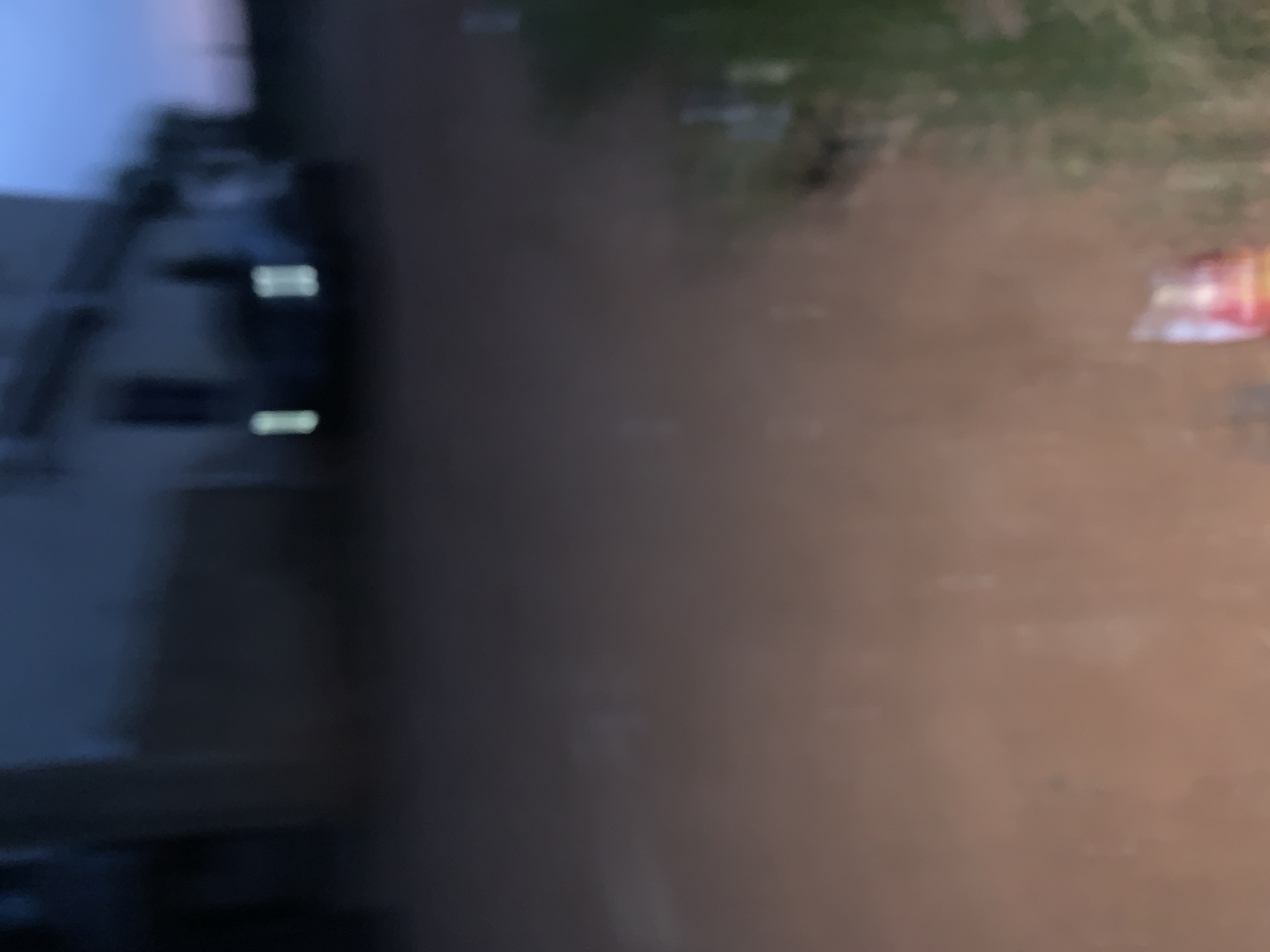 In the city of Bukuru, Nigeria, at dusk, a blurry image depicts a building with lights on, suggesting activity within. Adjacent to the building, a dark shape, possibly a vehicle, is faintly visible. The ground appears to be unpaved, with some scattered debris, including what looks like a crumpled red and white object in the foreground. The overall impression is one of a quiet, possibly residential or commercial area experiencing the transition from day to night. The lack of sharp detail makes definitive identification of specific objects and individuals impossible.