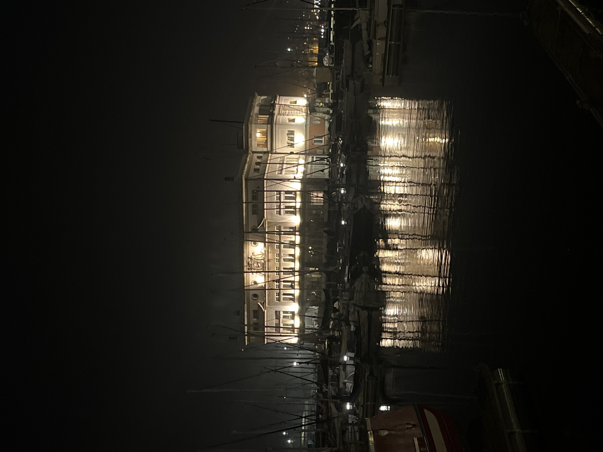At night, a long, three-story building with lit windows is reflected in dark, rippling water. Many sailboat masts are in the foreground, obscuring part of the building. Lights from the building reflect off the water. The building is in A Coruña, Spain.