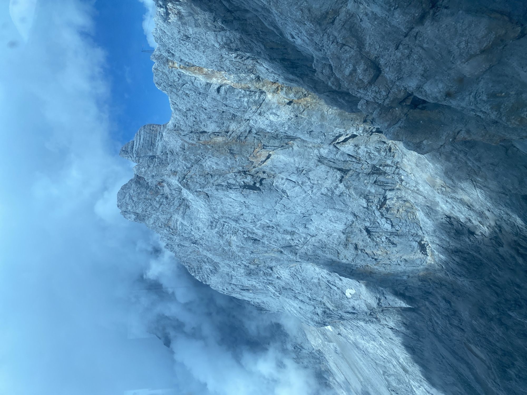 A jagged, imposing grey mountain face dominates the frame, its surfaces textured with countless fissures and ridges. Wisps of white cloud drift across the upper left portion of the image, obscuring the sky and parts of the mountain. In the distance, a sliver of bright blue sky is visible, hinting at clearer weather beyond the immediate cloud cover. A narrow, rusty-colored vein streaks down the center of the mountain face, contrasting with the predominantly grey rock. The lower section of the mountain is cast in deep shadow, creating a stark contrast with the sunlit upper sections. The overall impression is one of rugged, natural grandeur. This scene is likely a view of the Dachstein massif, which is accessible from Obertraun, Austria.