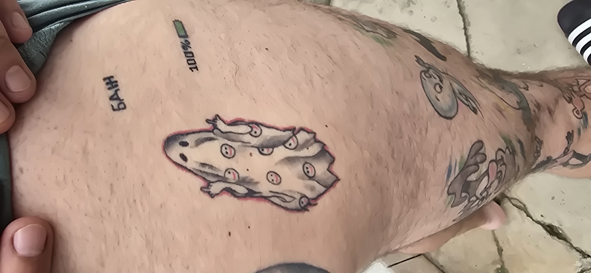 A close-up shot captures a person's upper leg and hip area, revealing an array of tattoos. A hand on the left gently pulls up green clothing, likely shorts or underwear, to expose the skin. The central and most prominent tattoo depicts a stylized, greyish-white, amorphous creature or ghost, dotted with numerous small, round faces featuring pink eyes. This design is outlined in a soft reddish hue.
Above this central tattoo, two smaller designs are visible: to the left, the Cyrillic word "БАН" is inked, and to the right, a fully charged battery icon displays "100%". The leg is further decorated with additional tattoos, including cartoon-like figures such as a grey oval with a face and a brown, possibly rabbit-like, character, extending across the thigh.
The setting appears to be outdoors in Govedare, Bulgaria, as suggested by the light-colored, cracked ground surface, which resembles concrete or large tiles, visible beneath the leg. A black and white striped item, possibly a sock or footwear, can be seen on the far right edge of the frame. The bright, even lighting indicates it is daytime. The image's focus is on showcasing the unique and whimsical collection of tattoos.