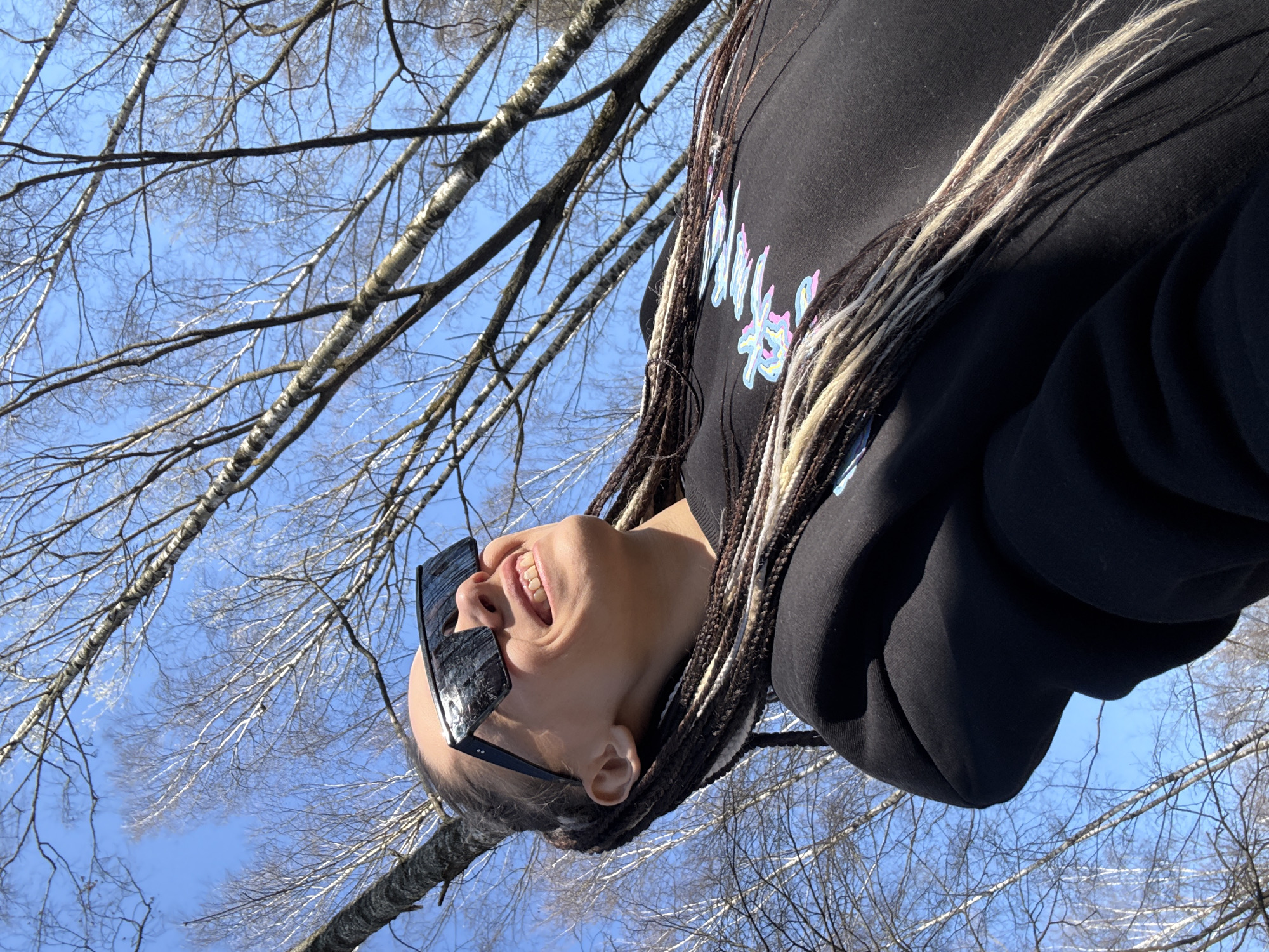 The image shows a person in a black hoodie and sunglasses, possibly of Asian descent, with braids in their hair, some of which are blonde, laughing and looking upwards. The person is positioned in front of a clear, blue sky with branches of leafless trees visible. The location is likely outdoors, in Unknown, Unknown, but specific city details are unclear.