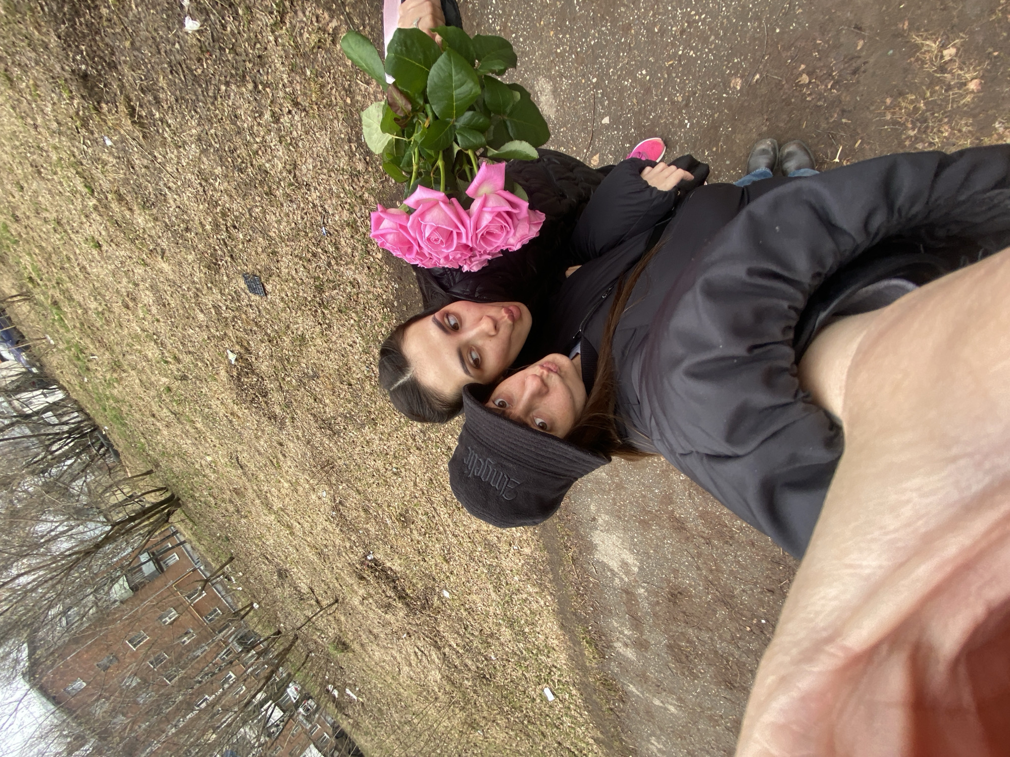This selfie, taken from a high angle, captures two young women posing outdoors in Votkinsk, Russia, during what appears to be late winter or early spring. The subjects are lying or sitting on the ground, looking up at the camera.
The woman on the left has long, dark hair and dark eyes, looking at the camera with a neutral expression. She holds a large bouquet of vibrant pink roses with prominent green foliage. She wears a dark jacket. Next to her, another woman in a dark jacket and a black bucket hat, which has white text appearing to read "Emgelic," looks directly at the camera with wide eyes. The arm and hand of the person holding the camera are visible in the foreground on the right.
The ground is covered with dry, light brown grass and bare patches of earth, indicative of a cold season. Scattered litter, including white paper scraps and a dark plastic object, is visible. In the background, through bare-branched trees, a multi-story brick building, possibly an apartment complex or institutional structure, can be seen. The overall lighting suggests an overcast day, and the heavy jackets worn by the women indicate cool weather. Pink sneakers are visible on the left, and dark boots or shoes are seen on the right. The image depicts a casual outdoor moment between the two women.