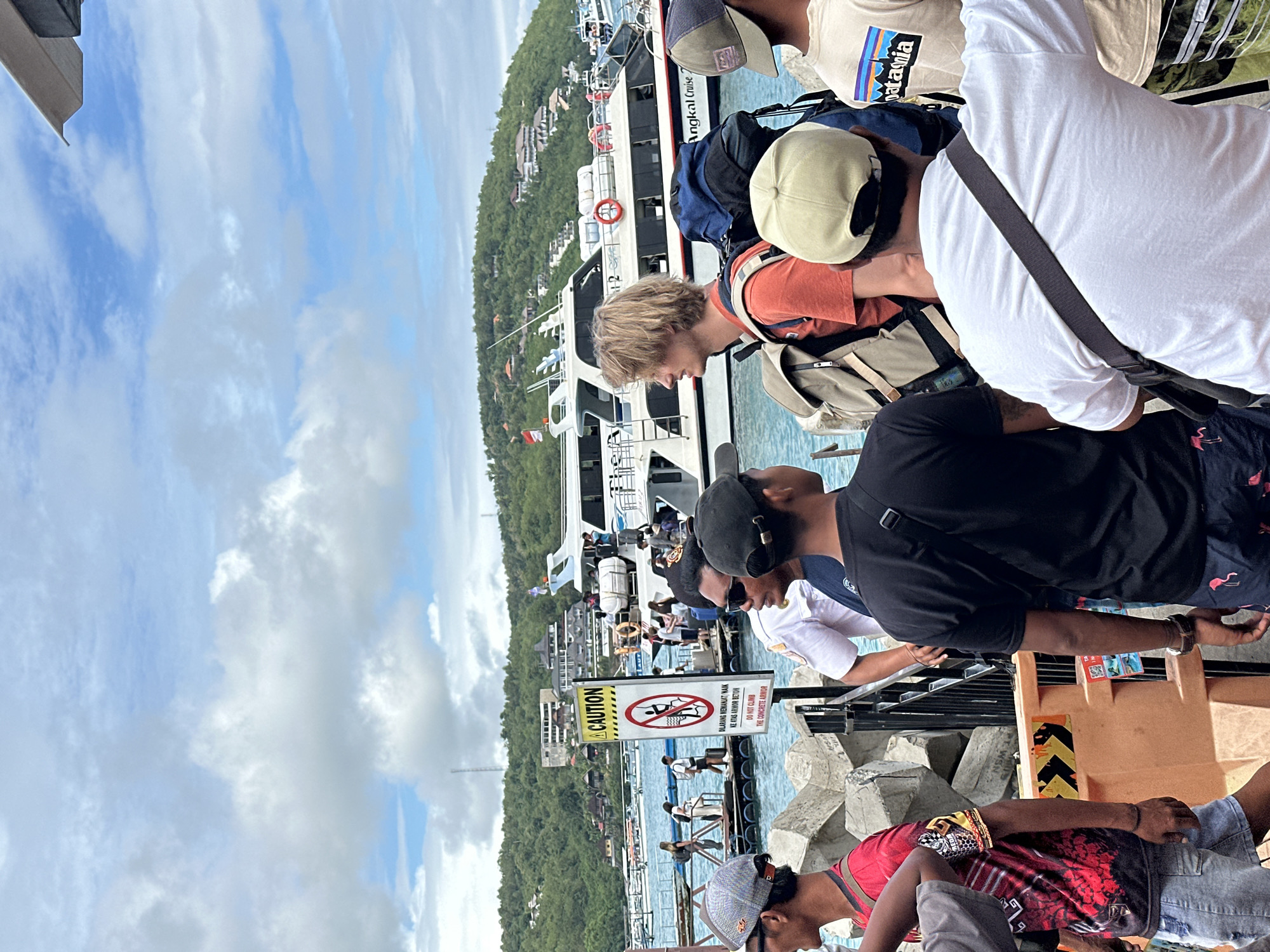 The image, taken in Nusa Penida, Indonesia, shows a group of people standing on a pier. A ferry boat is docked nearby. A caution sign in Indonesian and English warns against climbing the concrete armor. The sky is blue with scattered clouds, and green hills are visible in the background.