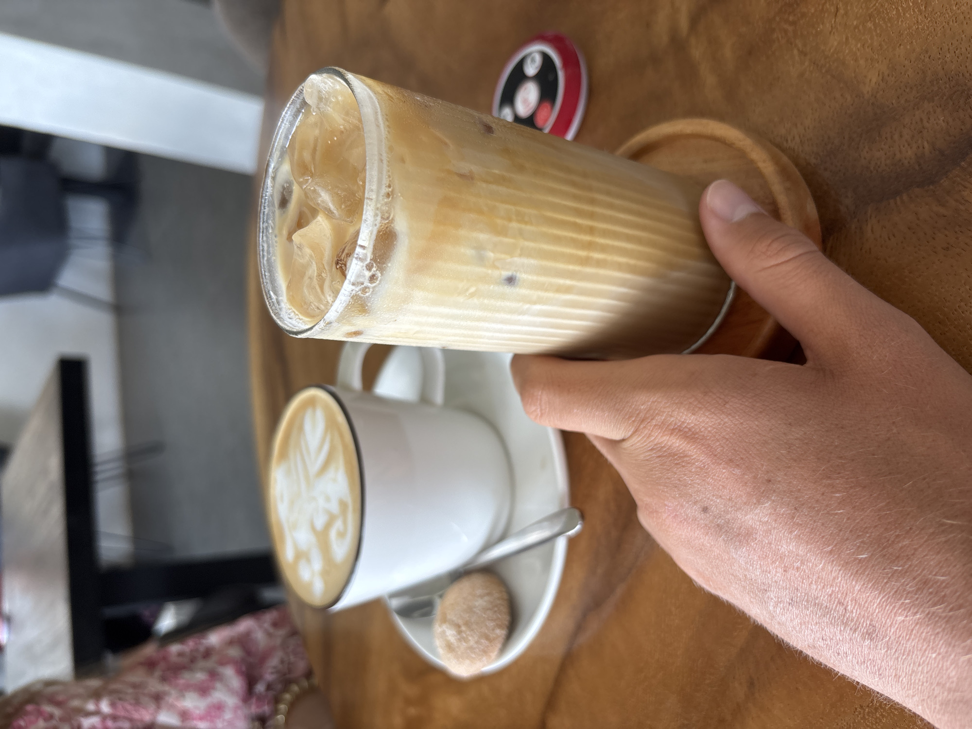 A person holds a glass of iced coffee, which is sitting on a small wooden coaster. Next to it is a white mug of latte with latte art, a spoon, and a small cookie, all on a white saucer. The table surface appears to be made of light-colored wood. The location is Kuta Selatan, Indonesia.