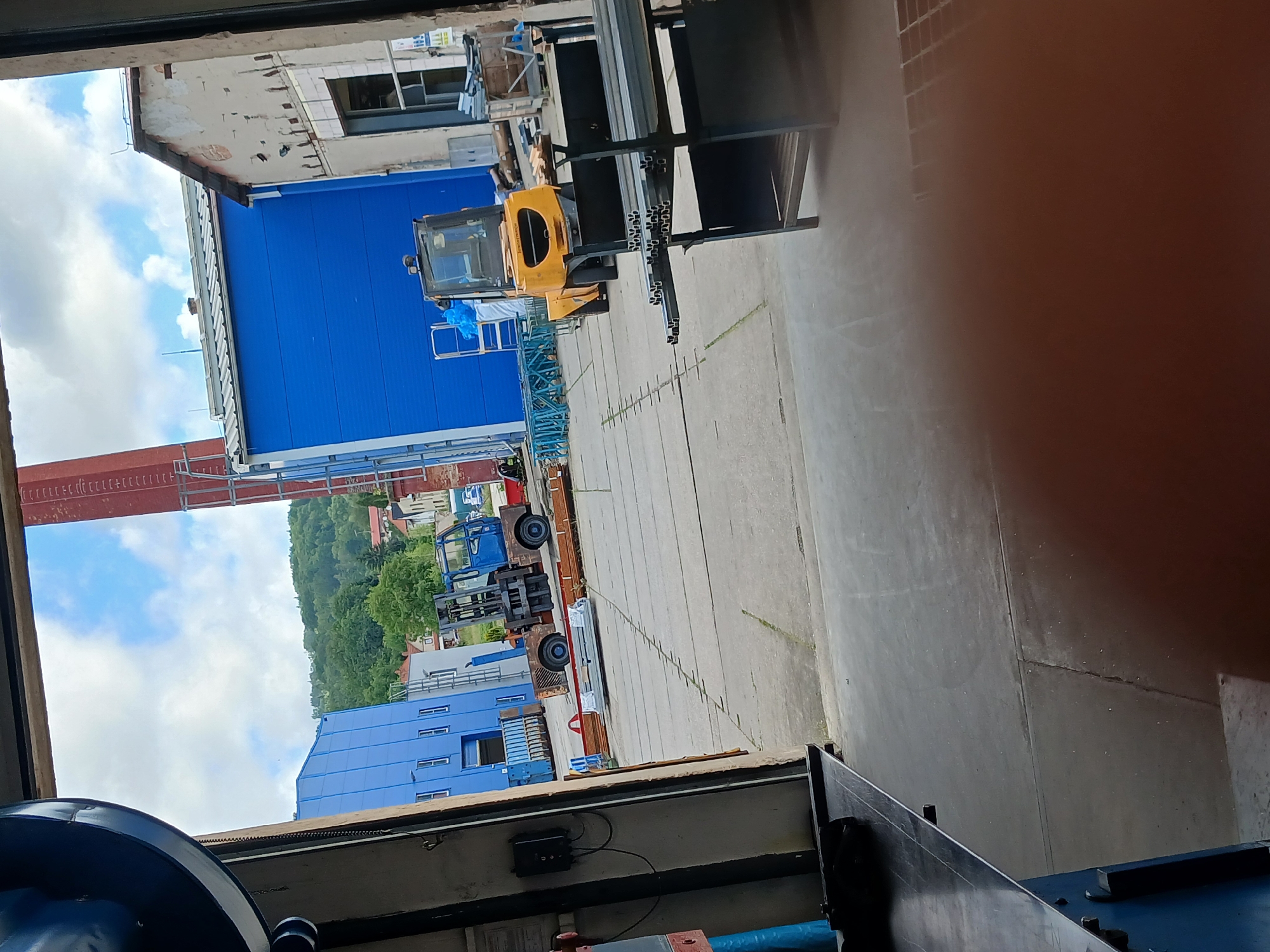 The image depicts an outdoor industrial area, likely a factory or construction site, viewed from within a building's open doorway. In the foreground, a metal table or workbench is visible on the left. The central area of the image shows a concrete yard with various industrial elements. A blue building with white-trimmed windows stands prominently in the middle ground. To its right, a small yellow forklift is parked. Further to the right, a more weathered, light-colored building with damaged facade is visible. In the background, a red brick chimney rises into a cloudy blue sky, and a lush green hill with scattered houses can be seen. The overall scene suggests a working environment with active construction or material handling operations. The context of Benešov nad Ploučnicí, Czechia, implies an industrial or commercial setting within that specific geographical location.