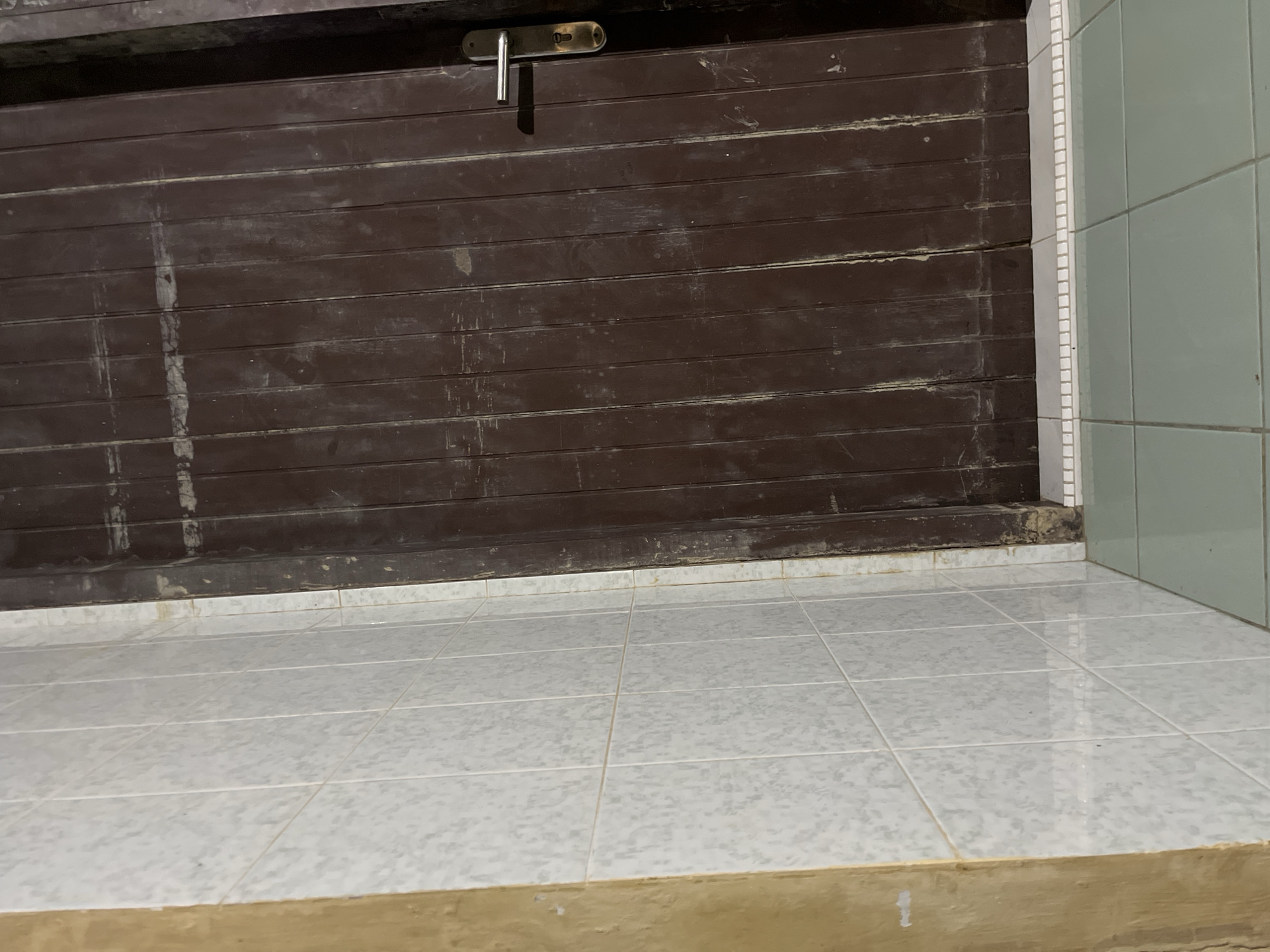 The image shows a section of a dark brown wooden door with vertical planks and a silver handle, positioned next to a wall covered in white textured tiles. The floor is tiled with light green squares. The scene appears to be indoors, possibly a hallway or room in a building. The lighting suggests it might be evening or a dimly lit interior. There are no people visible in the image. The location is stated to be in Kota Kuala Simpang, Indonesia.