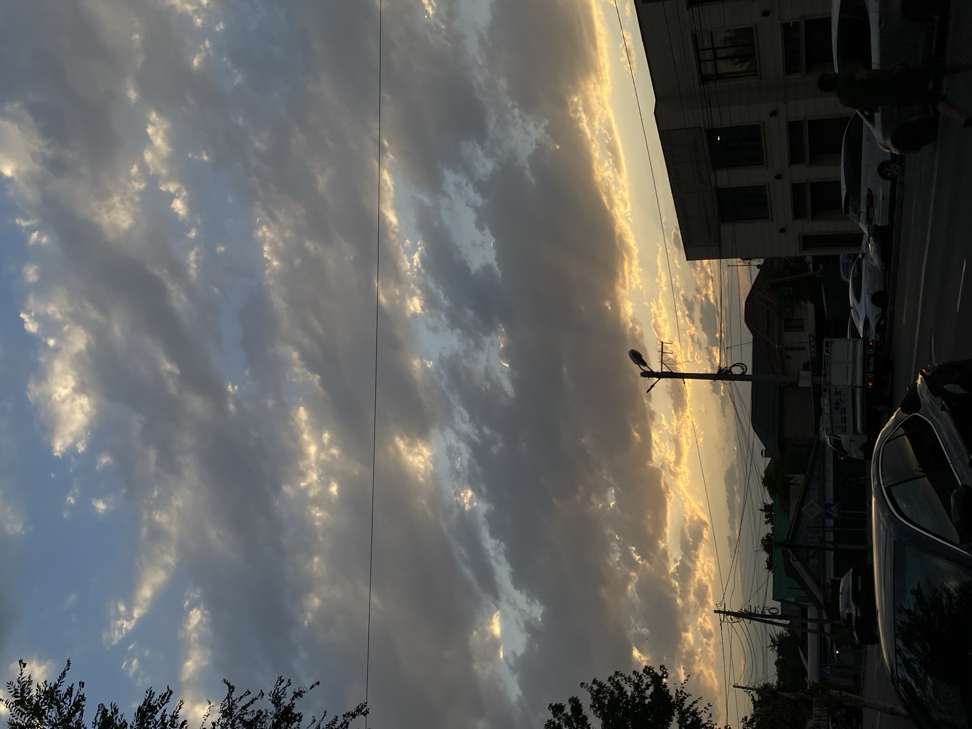 The image captures a street scene in Bishkek, Kyrgyzstan, during what appears to be either sunrise or sunset. The sky is filled with dramatic clouds, with patches of golden light breaking through, suggesting the sun is low on the horizon. Several power lines crisscross the frame, supported by utility poles, typical of urban infrastructure.
On the left side of the image, a dark-colored sedan is parked or moving slowly in the foreground, with its front end taking up a significant portion of the lower left quadrant. Further down the street, other vehicles are visible, including a white truck, a white sedan, and a white minivan. Buildings line the street; on the right, a multi-story building with large windows and a neutral-colored facade dominates the mid-right section. To the left of this building, residential houses with visible rooftops are present. A street lamp is also visible on the right side of the image.
The overall impression is a typical urban streetscape, with elements of nature, infrastructure, and human activity, set against a striking sky. No people are clearly identifiable or engaged in specific actions, though a person in a dark shirt and shorts can be seen walking on the sidewalk on the right side of the image.