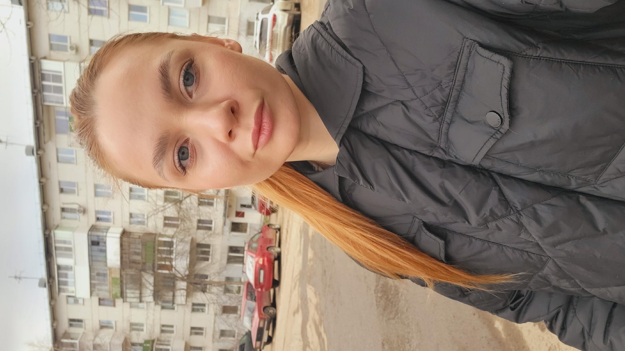 A selfie depicts a light-skinned woman with light blonde hair and blue eyes, positioned outdoors. She's wearing a black quilted jacket. The background reveals an urban environment with a multi-story building and parked cars, typical of a residential area. Location possibly in Chaikovsky, Russia.