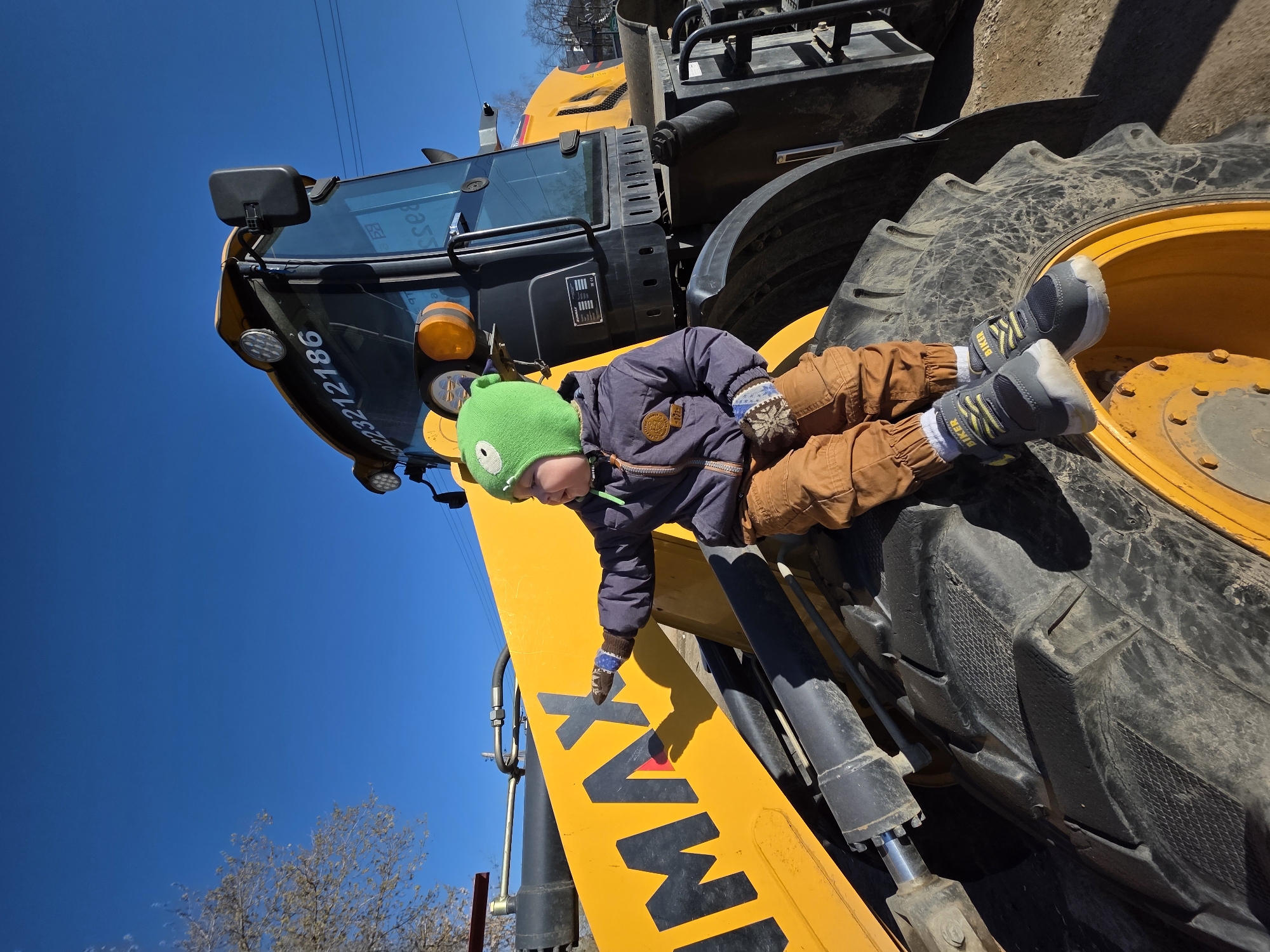 A low-angle shot shows a child lying on a yellow tractor. The child is wearing a green hat, blue jacket, and brown pants, and has on grey and yellow sneakers. The child's right hand is pointing towards the word "MAX" printed in bold, black letters on the yellow tractor arm. The tractor has a large black tire, and a dark blue sky is visible in the background. The tractor has the number 92233-12186 written on it. The location is the city of Chaikovsky, Russia.