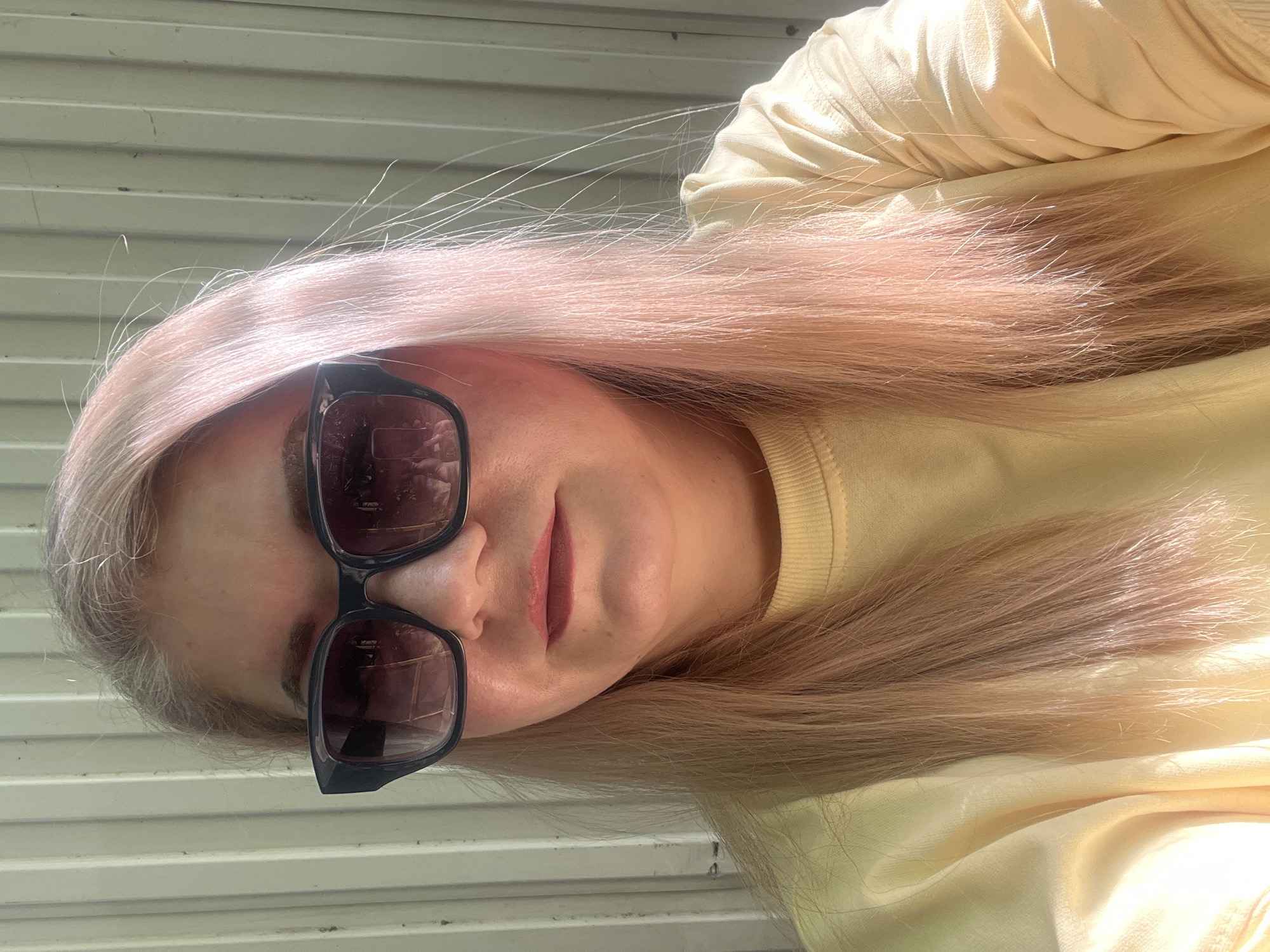 A person with long, light pink hair and sunglasses poses for a selfie. They are wearing a pale yellow sweater. The background appears to be an exterior wall with horizontal siding. There is no information to establish the location as Izhevsk, Russia.