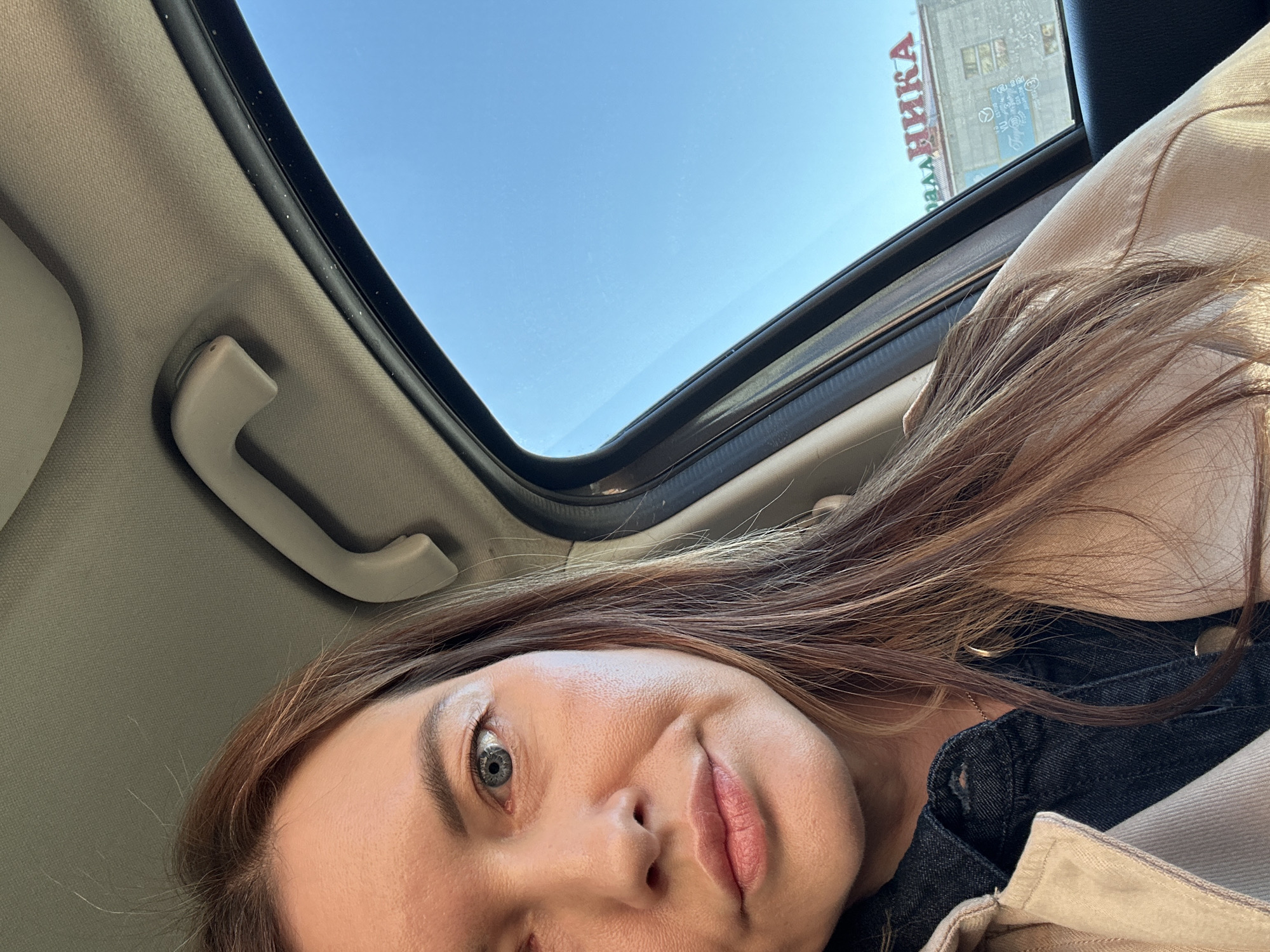 A woman with light skin, brown hair, and gray eyes is visible in a selfie taken inside a car. She is wearing a beige jacket over a dark denim top. The car's sunroof is open, revealing a clear, blue sky. Through the sunroof, a building with a sign in Cyrillic script, "ДЕТСКАЯ КНИГА" (meaning "CHILDREN'S BOOK"), is partially visible, indicating a location in the city of Chaikovsky, Russia. An interior car handle is visible on the upper left.