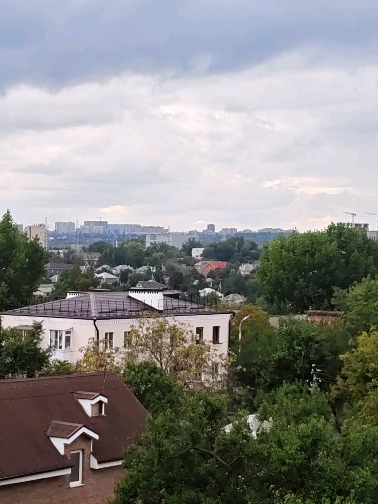 This is an aerial, eye-level photograph taken on an overcast day, looking across a suburban landscape towards a distant city skyline. The foreground is dominated by the roofs of several buildings, with a large, brown, corrugated metal roof on the lower left, featuring two dormer windows with white trim. Behind this, a cream-colored building with a white decorative cornice and numerous windows is visible. Lush green trees are interspersed throughout the mid-ground, partially obscuring the view of smaller houses with red tile roofs. In the distance, a sprawling urban area is discernible, characterized by numerous apartment buildings and construction cranes, indicating ongoing development. The sky is filled with dense, grey clouds. The general impression is of a temperate climate, with abundant vegetation and a mix of residential and high-rise construction typical of a growing city. The specific location is the Voroshilovsky district of Rostov-on-Don, Russia.