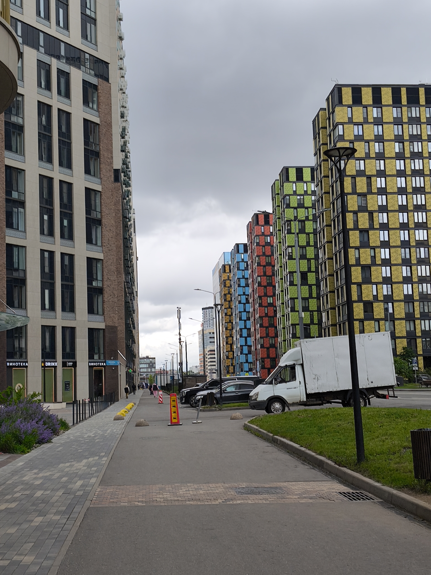 The image shows a street in Moscow, Russia, lined with modern, multi-story apartment buildings of varied colors and architectural styles. The buildings are tall, with repeating window patterns. A white delivery truck is parked on the side of the road, partially obscuring a row of colorful buildings. Sidewalks are present on both sides of the street, with landscaping and pedestrian barriers. People are seen walking in the distance. The sky is overcast, suggesting a cloudy day. The scene is urban and functional, capturing a typical street view in a modern Russian city.