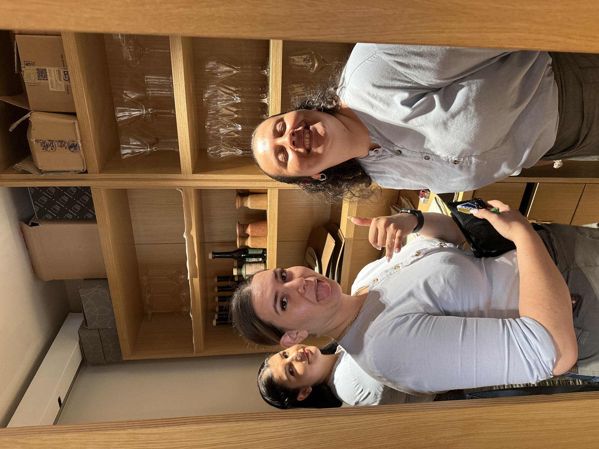 Three women stand in a narrow hallway with wooden shelving units filled with glassware and bottles. The woman on the right is laughing with her eyes closed and her head tilted back. She is wearing a light blue button-up shirt and an apron. The woman in the middle is sticking her tongue out at the camera and giving a thumbs up. She is also wearing a light blue button-up shirt and an apron. The woman on the left is looking at the camera with a slight smile. She is wearing a light blue shirt. The shelving units appear to be in a restaurant or bar. The image is taken in Nicosia, Cyprus.