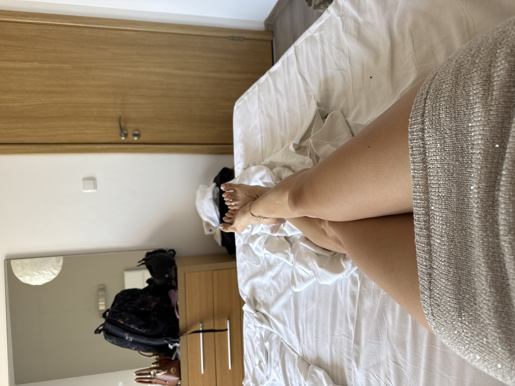 A person's legs, adorned with a sparkling skirt and a beaded anklet, are crossed on a white, rumpled bed in a hotel room in Sozopol, Bulgaria. The room features a wooden dresser with bags and a mirror, a white duvet, and a wooden door.