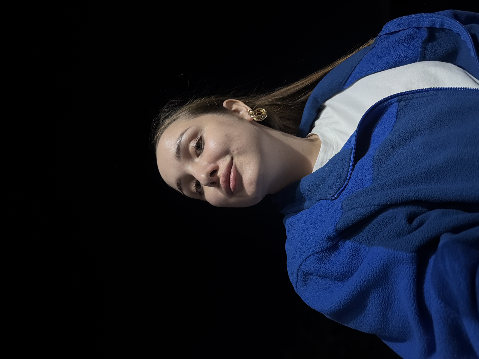 A low-angle shot portrays a young woman with light skin, brown hair, and a slight smile, positioned against a stark black background. She is wearing a white t-shirt underneath a blue fleece jacket with a zipper. A gold earring is visible on her left ear. The scene's location within the city of Chaikovsky, Russia, cannot be determined from the provided image.
