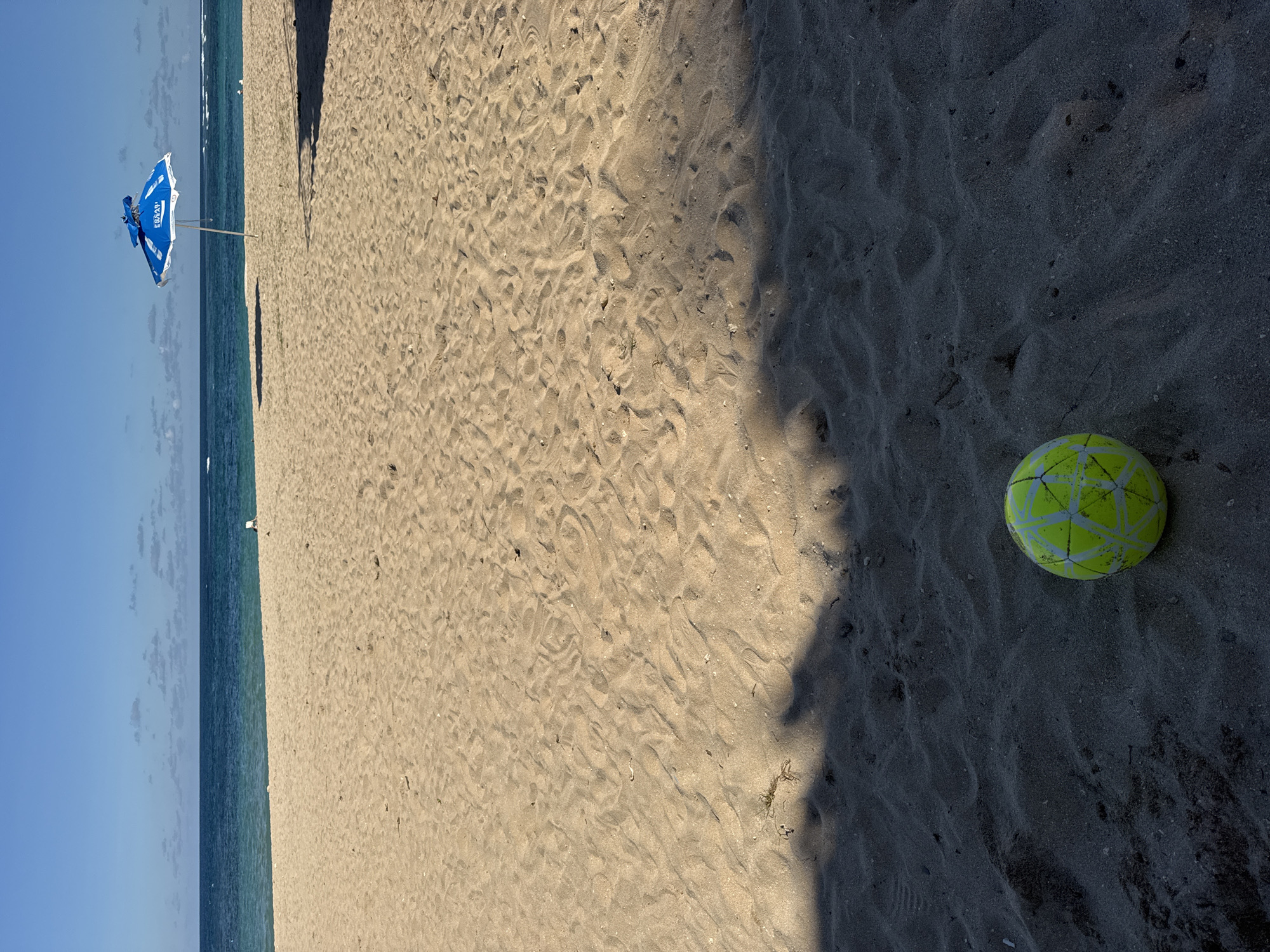 A horizontal, eye-level shot captures a beach scene, with the sky and ocean on the left and sand occupying most of the frame. A blue and white beach umbrella stands near the shoreline. A shadow stretches across the right side of the image, partially covering a yellow soccer ball resting on the dark sand. The beach's sand is varied in texture and color, from light tan to dark grey.