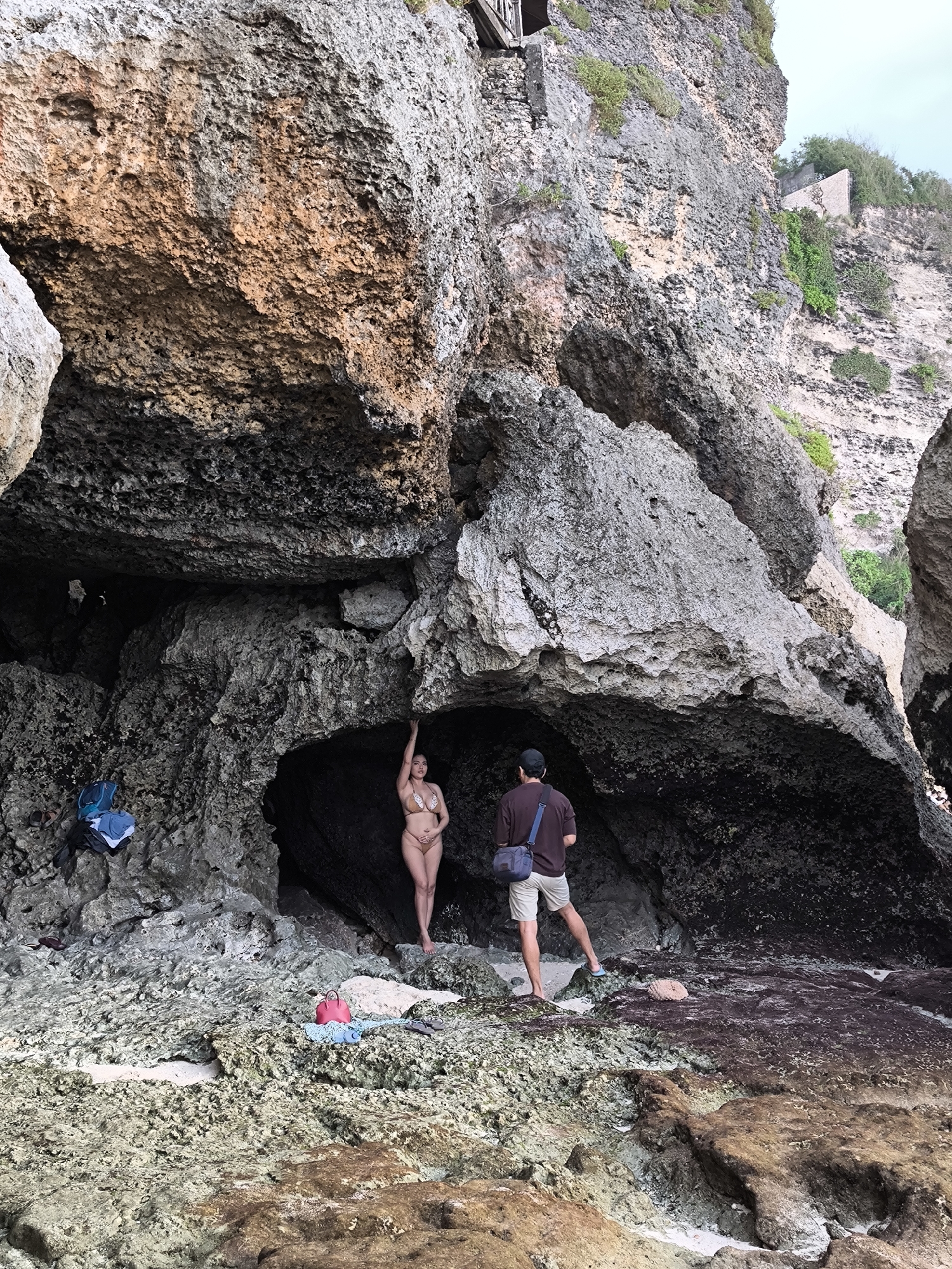 A medium shot depicts a woman in a bikini posing inside a cave while a man stands nearby, presumably taking a photo of her at Uluwatu, Indonesia.
The woman, light-skinned, is positioned in the center of the cave. She has one arm raised, holding onto the cave ceiling for support. The man, standing on the right, is wearing a dark shirt, khaki shorts, a baseball cap, and a shoulder bag. He is looking towards the woman, appearing to be taking a picture.
The cave's entrance is surrounded by large, rugged rocks. The beach in front of the cave is rocky, with some sand patches. A blue backpack and some clothing are on the rocks to the left of the woman.