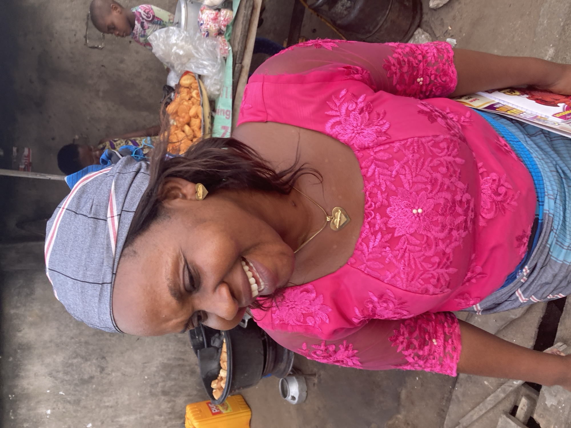 The image shows a woman smiling at the camera, wearing a pink lace top, a grey headscarf, and a heart-shaped necklace and earrings. She is standing near a pot of fried food and a yellow container labeled "Purd". In the background, other people and goods are visible. The location is likely a street-side food stall in Aba, Nigeria.