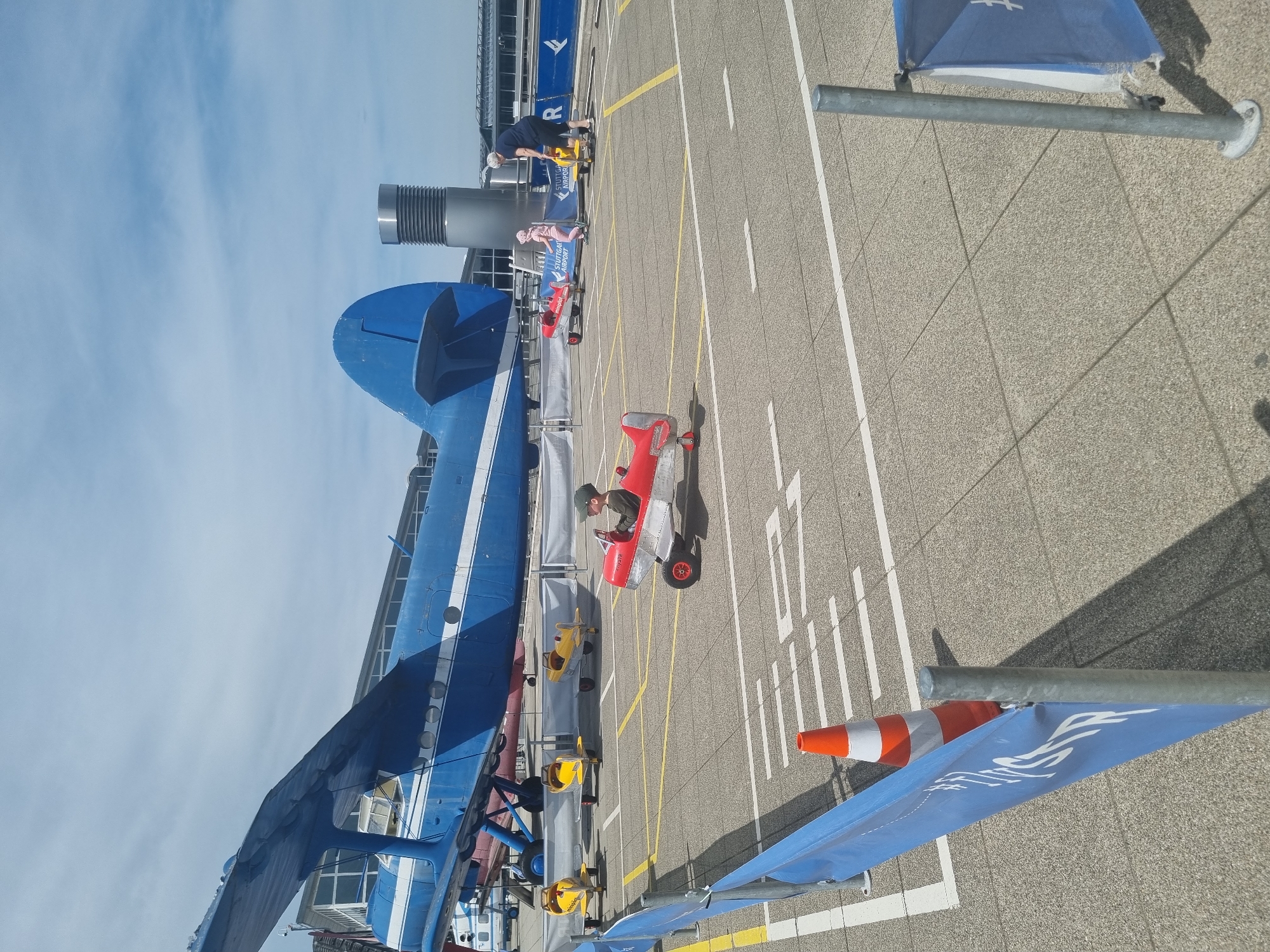 The image shows an outdoor scene at Stuttgart Airport in Echterdingen, Germany. A large blue and white biplane dominates the left side of the frame. In the center, a child wearing a green hat and jacket sits in a small red airplane toy. Several other airplane toys, primarily yellow, are parked nearby. An adult in a dark top is standing next to the biplane on the left. The ground is marked with yellow and white lines, suggesting a parking area or runway. A blue flag on a pole is visible in the foreground, and an orange traffic cone stands near it. In the background, airport buildings are visible. The sky is blue with scattered clouds.