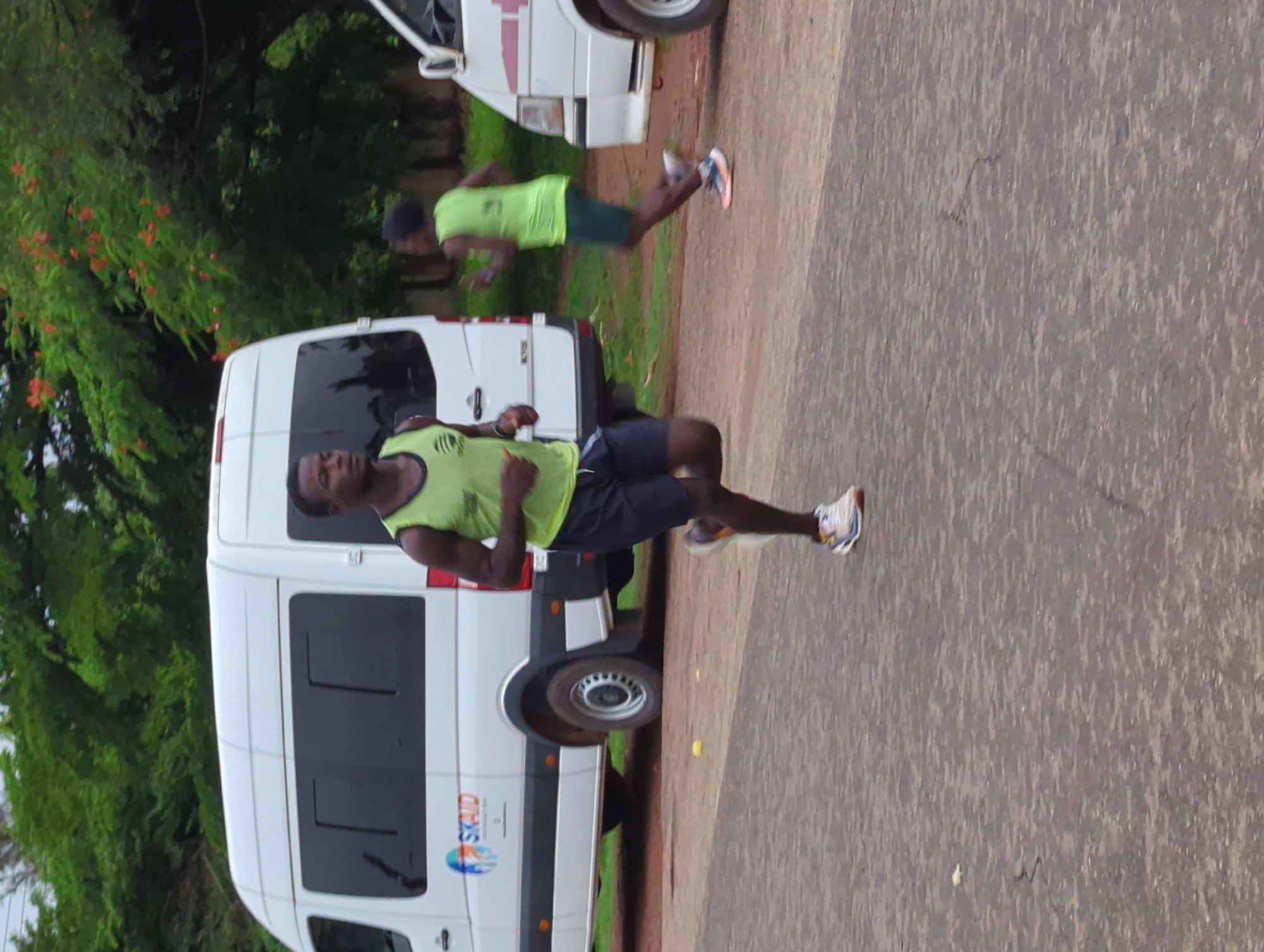 Two male runners are jogging on a paved surface in Gidan Waya, Nigeria. Both are wearing athletic attire with bright yellow tank tops and dark shorts. They appear to be pacing each other, running side-by-side. A white van is parked to their left, showing an emblem. The background includes green trees and plants.