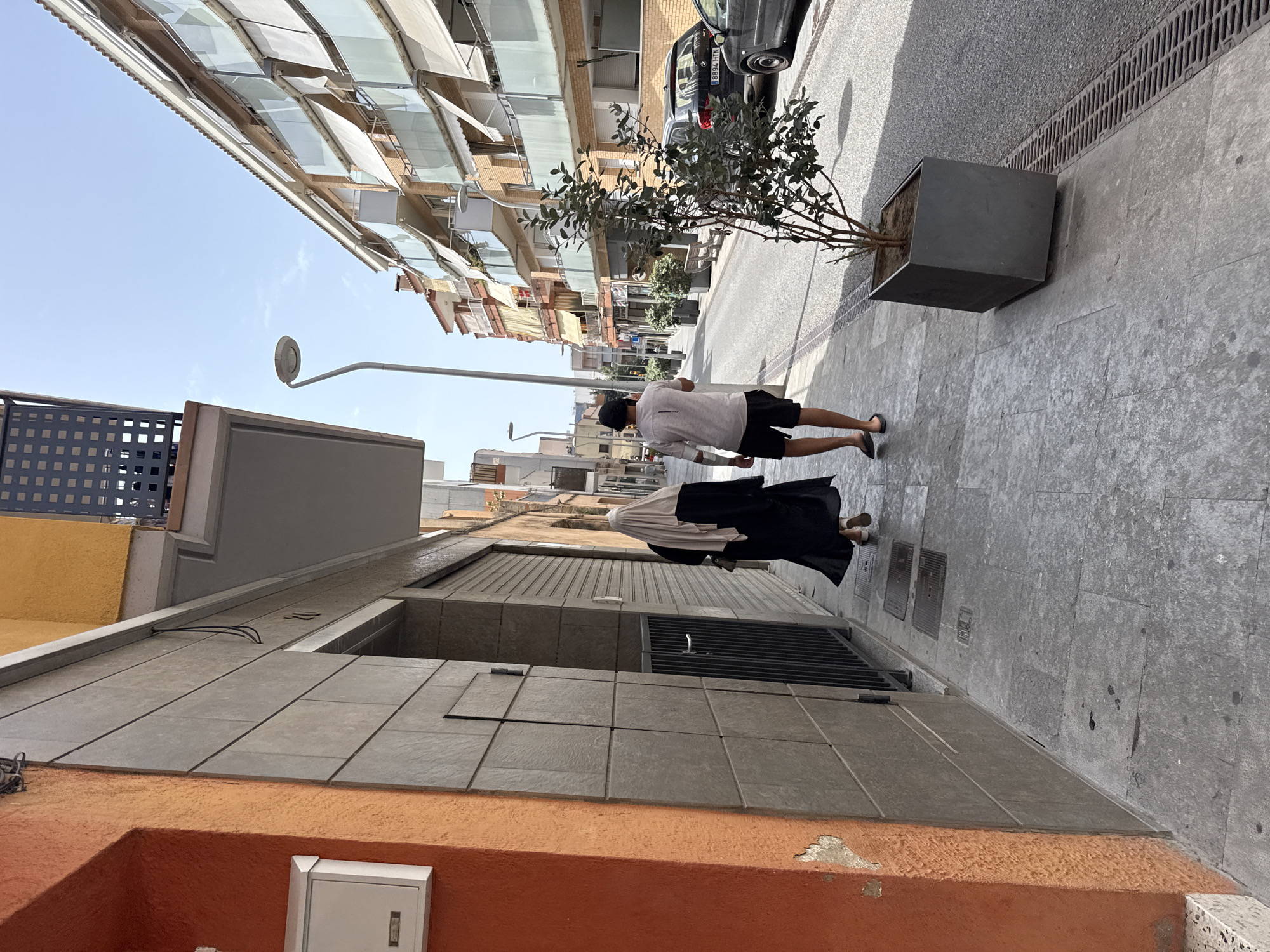 In Blanes, Spain, two individuals walk along a sunlit city street. On the left, a person is partially visible from behind, wearing a black abaya and a beige hijab. They are walking away from the camera, towards the right side of the frame. To their right, a man wearing a light-colored t-shirt, black shorts, and flip-flops also walks away from the camera. The street is lined with buildings, featuring balconies with awnings. A parked car with a license plate reading "8894 HTN" is visible on the right side of the street. A dark gray planter with a small tree sits on the sidewalk. The pavement is made of grey flagstones.