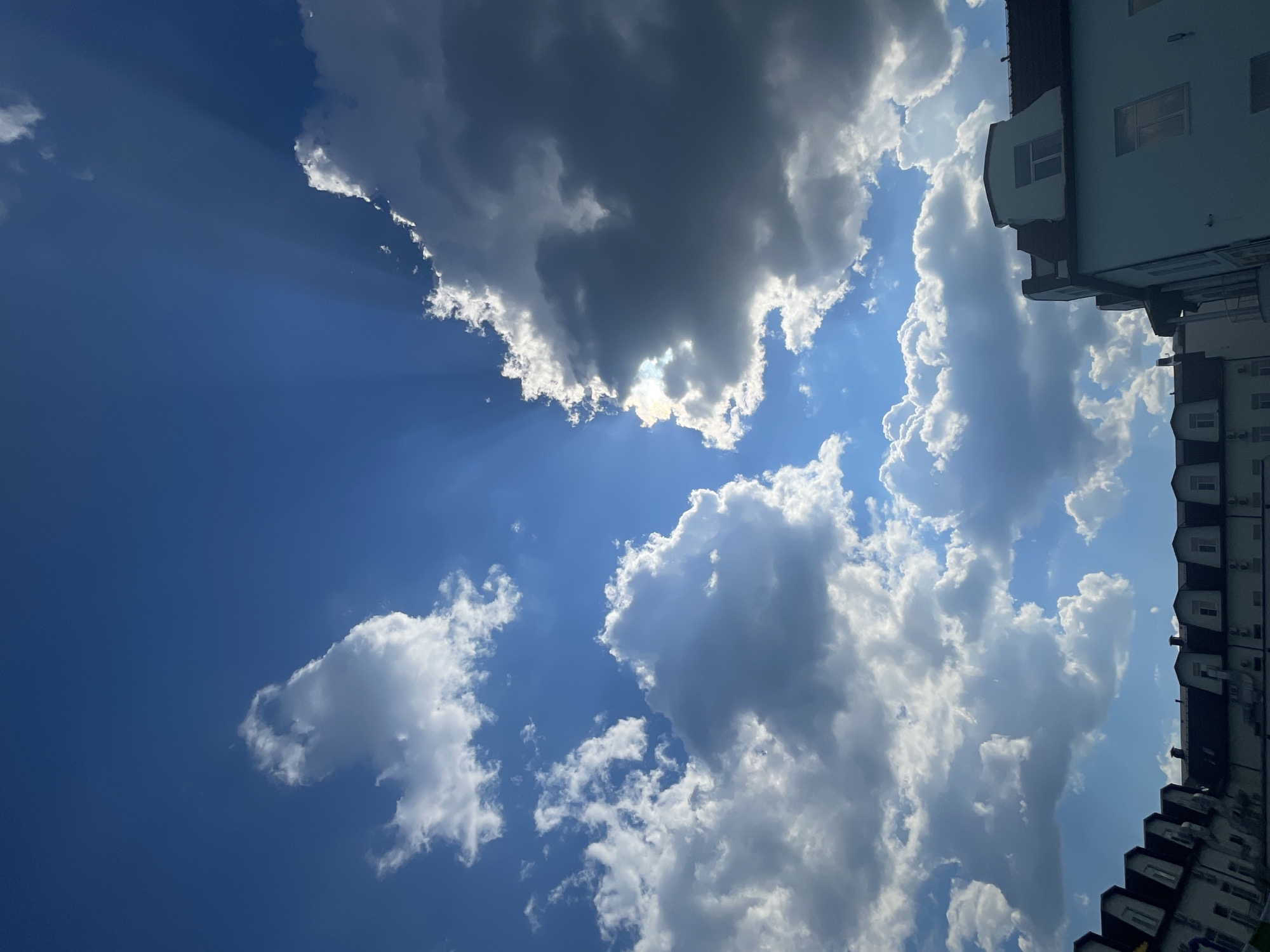 The image depicts a bright, partly cloudy sky over a city. The sun is visible behind a large, dark cloud, with rays of light emanating from it, creating a dramatic effect. The sky is a vibrant blue, with fluffy white clouds scattered throughout. In the foreground, the rooftops of several buildings are visible, suggesting an urban setting. The architecture appears to be of a traditional European style, with dormer windows and pitched roofs. The lighting suggests it is daytime, likely late morning or early afternoon, given the angle of the sun's rays. The scene is devoid of any people. The location is identified as Pristannoye, Russia.