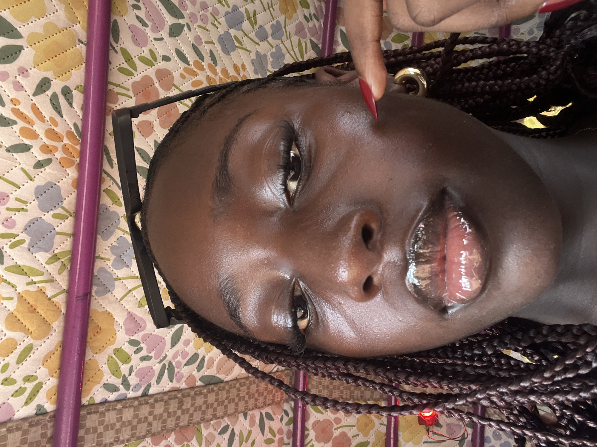 A close-up, slightly angled view of a dark-skinned person with braided hair. A finger with a red nail touches their cheek near the ear, which has a gold hoop earring. The person has glossy lips. Glasses are positioned near their face, and the background consists of a floral patterned surface with a purple object. The location is not identifiable.