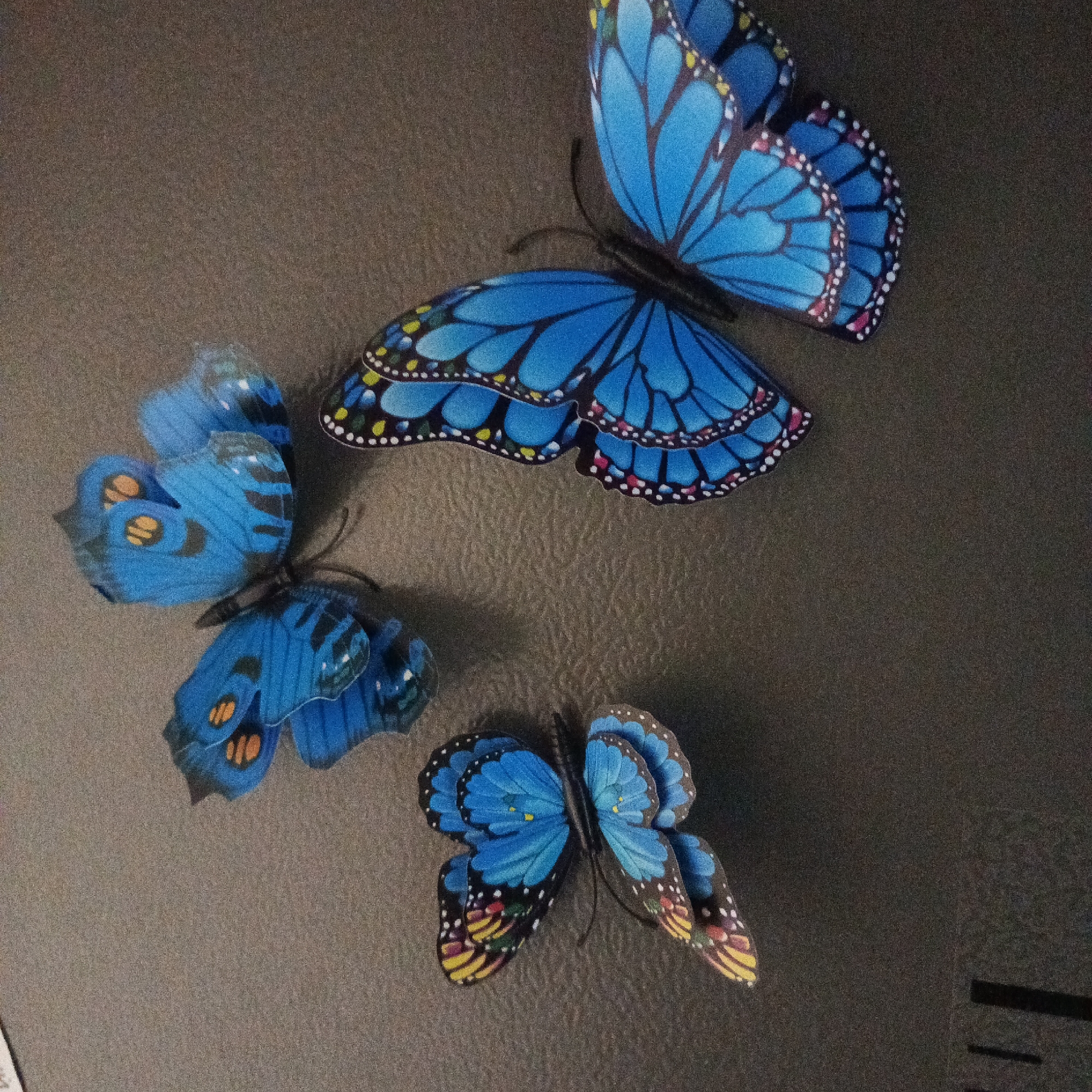 The image shows three blue butterfly decorations affixed to a dark, textured surface. The largest butterfly is positioned in the upper right, the second largest in the lower center, and the smallest in the upper left. The butterflies have varied patterns and wing shapes. The background is a dark grey or brown color with a visible texture. The scene is indoors. There is no information to determine the location of the image as Марковский, Russia.
