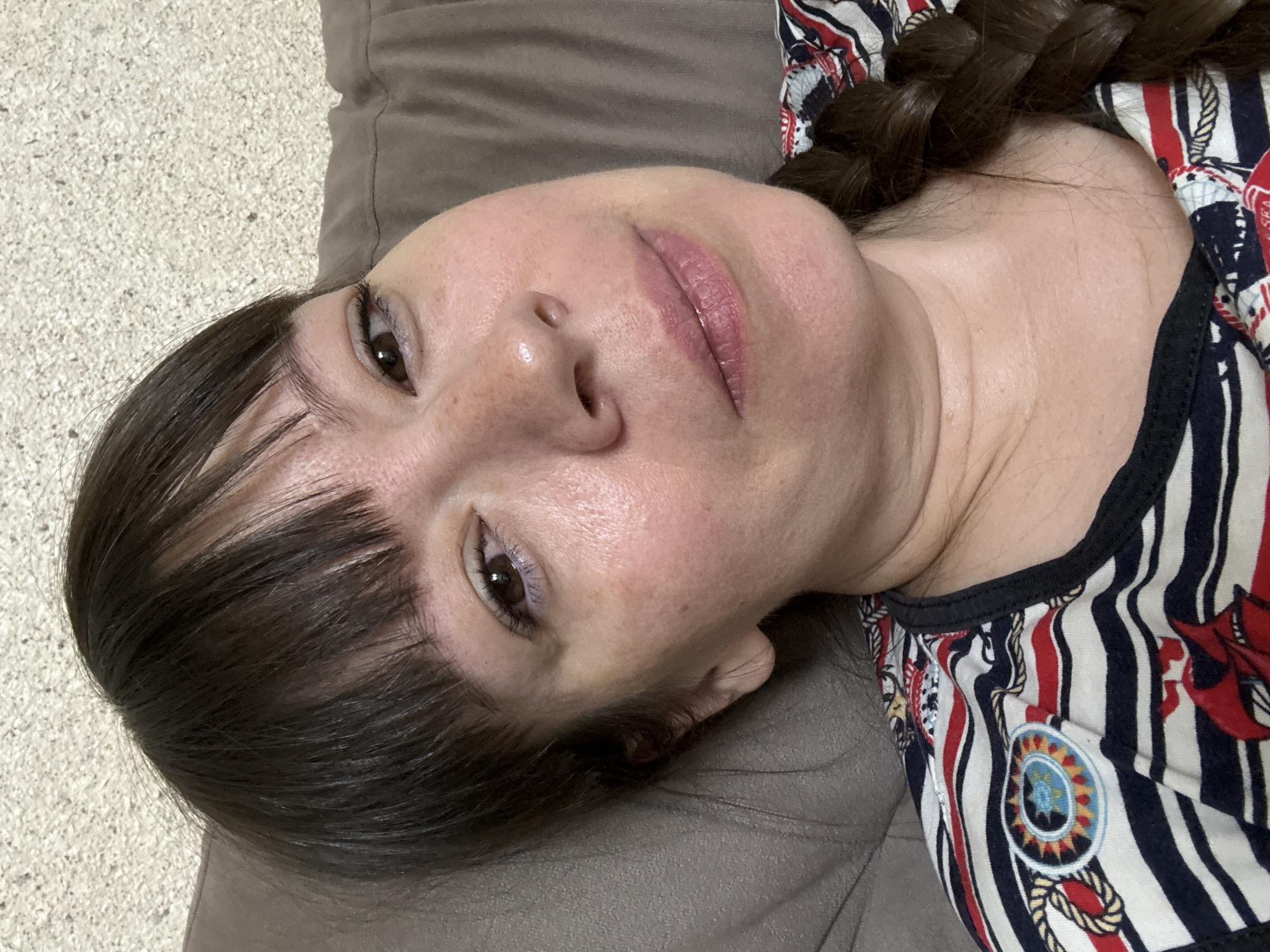 A close-up selfie shows a woman with dark hair and bangs lying down, with her head resting on a brown cushioned surface. She is wearing a striped shirt with nautical motifs, including a ship and rope designs. Her right eye is visible, along with her nose and mouth. The background consists of a textured, light-colored wall and the brown cushion. The image appears to be taken indoors.
